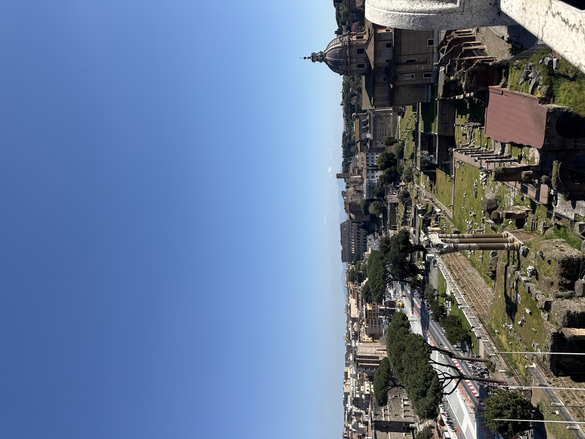 The image shows a cityscape of Rome, Italy. Landmarks include the Colosseum and the Roman Forum, visible amongst other buildings and green spaces. A road with traffic and pedestrians runs adjacent to the Forum. In the background, a large dome, likely part of a prominent church, is visible. The sky is a clear, bright blue.