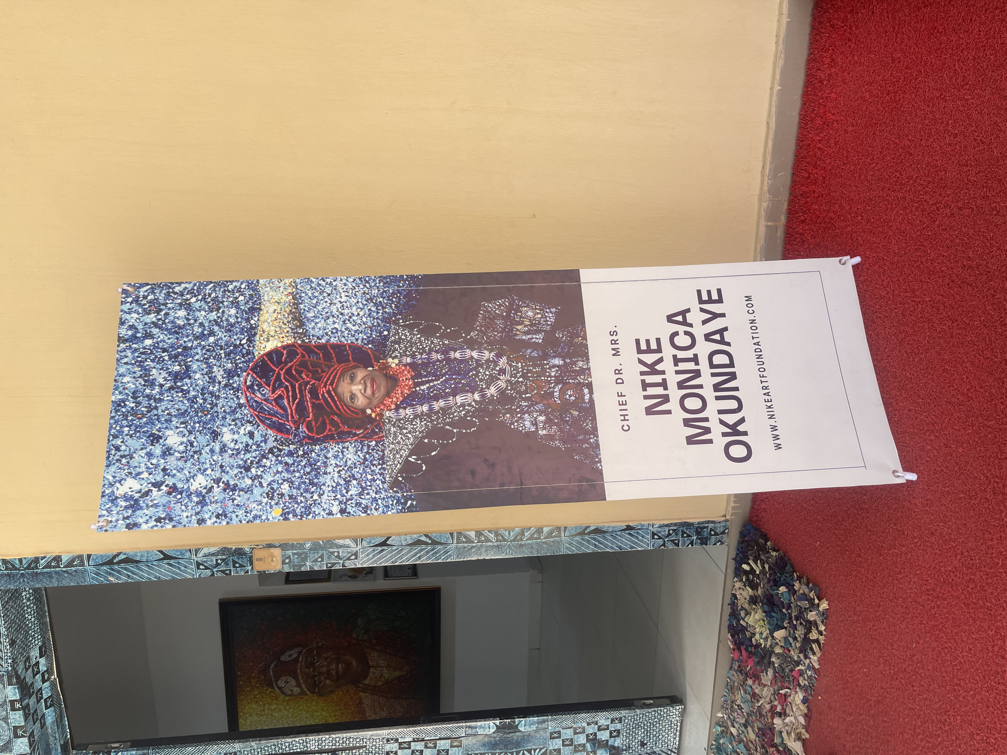 The image captures a vertical banner displayed at an entrance. The banner features a portrait of a woman, identified by accompanying text as Chief Dr. Mrs. Nike Monica Okundaye. She is depicted wearing traditional red beaded headwear and a necklace, with a patterned top. The background behind her in the portrait is a mosaic of blues, whites, and darker tones.
To the right of her portrait, white text against a dark grey background reads "CHIEF DR. MRS." above her name "NIKE MONICA OKUNDAYE" in large, bold letters. Below her name, the website "WWW.NIKEARTFOUNDATION.COM" is visible.
The banner is positioned upright, secured at its top and bottom corners, and stands against a plain light beige wall on the left. On the right, a textured red carpet covers the floor. Below the banner, at the entrance threshold, a colorful, shaggy rag rug in shades of blue, white, and red is visible next to the red carpet. Further inside the building, a large framed artwork depicting a face in warm tones can be seen hanging on a wall, suggesting an art gallery or cultural space. Decorative blue and white patterned tiles or fabric adorn the lower part of the doorway/wall behind the banner. The overall scene suggests a public entrance, possibly to an art foundation or exhibition in Abuja, Nigeria.