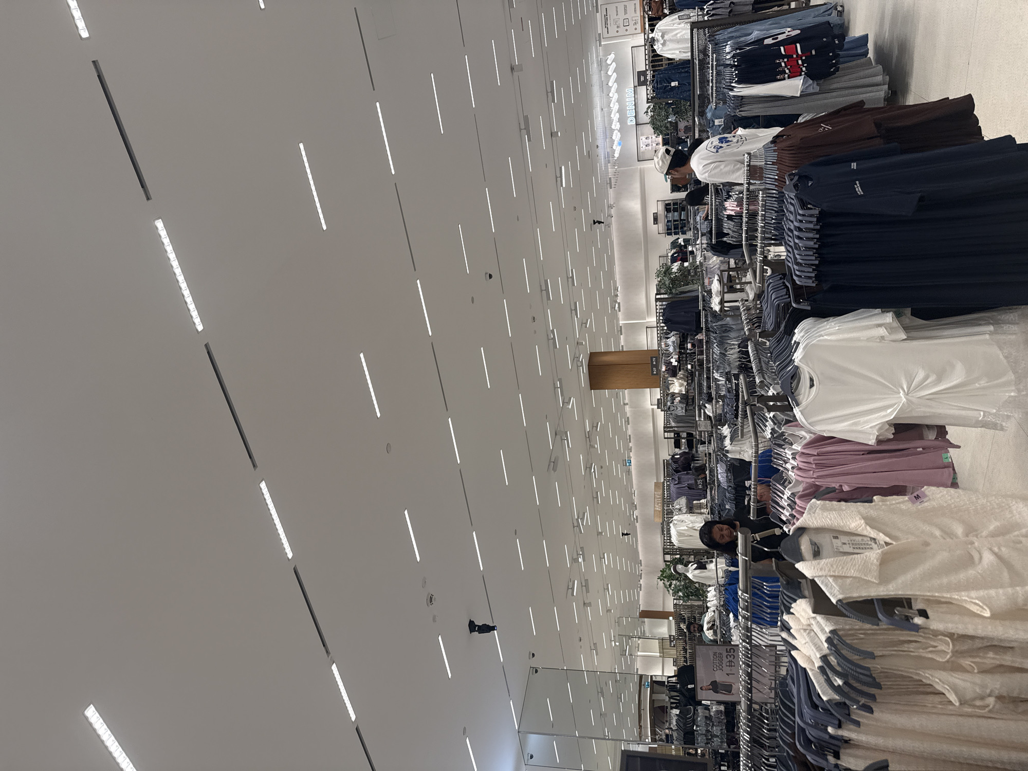The image depicts the interior of a spacious, well-lit clothing retail store. The ceiling is adorned with numerous long, linear LED light fixtures arranged in a grid pattern, creating a bright and modern ambiance. Rows of clothing racks are meticulously organized throughout the store, displaying a variety of apparel such as t-shirts, dresses, and possibly activewear, in various colors and styles. Some garments are displayed on mannequins, while others are hung on hangers.
In the foreground, several racks of clothing are visible, with a focus on lighter-colored tops and dresses. Further back, the store extends into a larger retail space with more racks and displays. There are several individuals present in the store. One woman with dark hair and wearing a black top is seen browsing near a clothing rack in the middle ground. A person wearing a white cap and a white t-shirt is visible in the right-middle ground, facing away from the camera. Another person, a man with dark hair and a beard, is partially visible in the left-middle ground, standing near a display.
There are also signs visible in the store, including one that reads "DENIM" in illuminated letters against a dark background, suggesting a dedicated section for denim products. Another sign indicates "SMART FITTING ROOMS". The store appears to be in a modern shopping mall environment, characterized by its clean design, ample lighting, and organized merchandise. The scene is consistent with a retail clothing store located within a city like Dubai, United Arab Emirates, which is known for its numerous large and contemporary shopping centers.