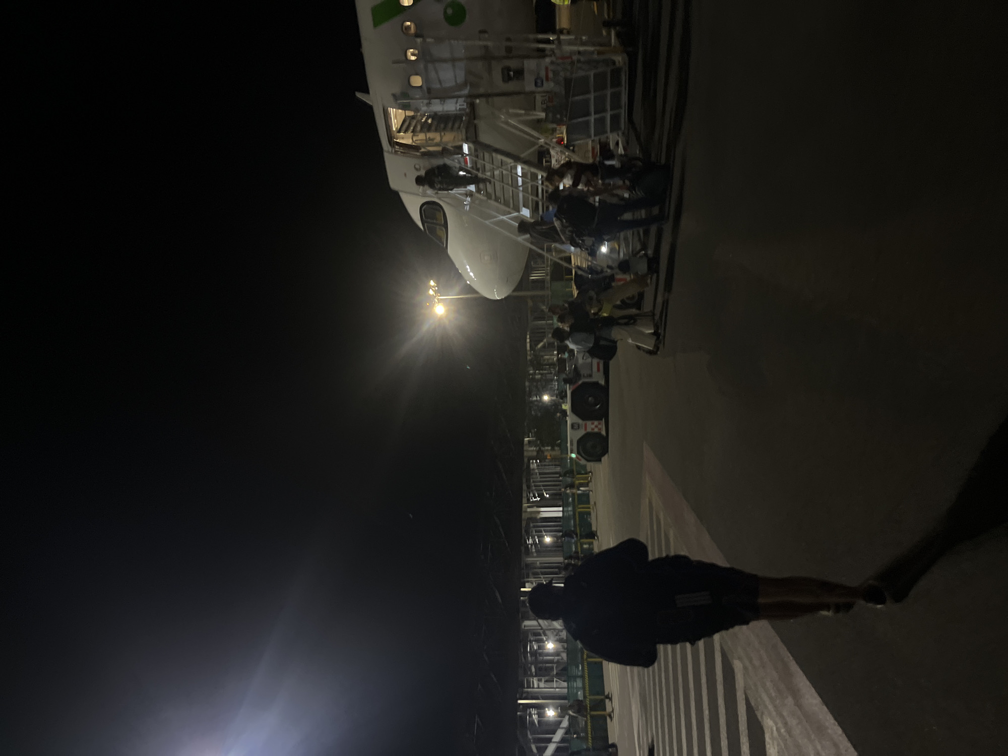 At night in Puerto Escondido, Mexico, an airplane is parked on the tarmac, with passengers disembarking via a mobile staircase. A person is walking toward the aircraft. The area is illuminated by overhead lighting.