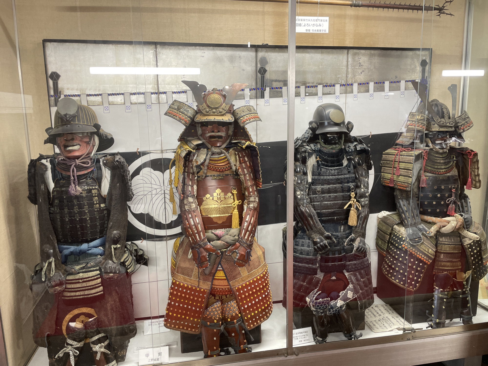 This image displays a museum exhibit in Iga, Japan, featuring four samurai armors, each set in a glass display case. The armors are positioned from left to right, with the first one having a red face mask and a dark helmet with a stylized crest. The second armor is a vibrant red and gold ensemble with a prominent chest plate and elaborate leg coverings. The third armor is primarily black, with a dark helmet and intricate detailing on the chest and arms. The fourth armor, partially visible on the far right, also appears to be dark with red and gold accents. The background of the exhibit consists of a large screen with a black and white circular motif and horizontal bands of blue and white patterns, suggesting a historical or cultural theme. Various labels and descriptive texts are placed around the display, providing information about the artifacts. The lighting within the exhibit is bright, illuminating the armors and their intricate craftsmanship.