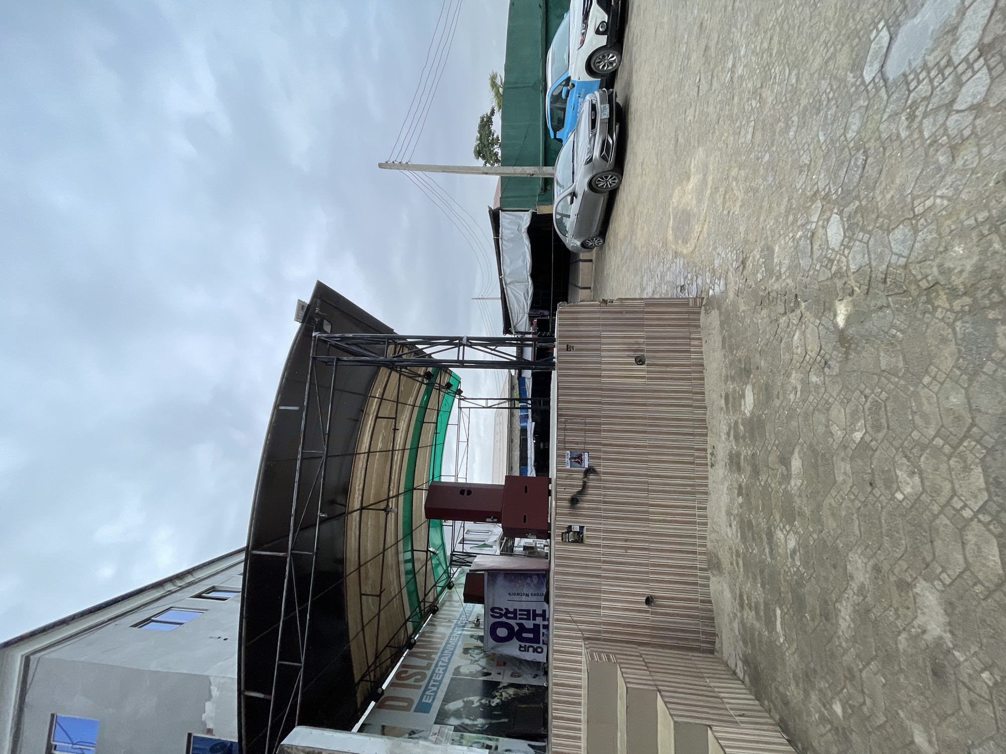 The image depicts an outdoor stage area in Ado, Nigeria, under a cloudy sky. The stage is covered by a canopy supported by metal poles. Behind the stage, a banner reads "D ISLAND YOUTH ENTERTAINMENT CENTER". On the stage, there are speakers and a large, red amplifier. To the right of the stage, there is a paved area with two cars parked: a silver sedan and a white SUV with a blue door. The ground in front of the stage appears to be wet and shows a patterned pavement. The overall scene suggests an event or entertainment venue, possibly with a history of use, given the weathered appearance of the pavement and the slightly worn condition of the stage cover.