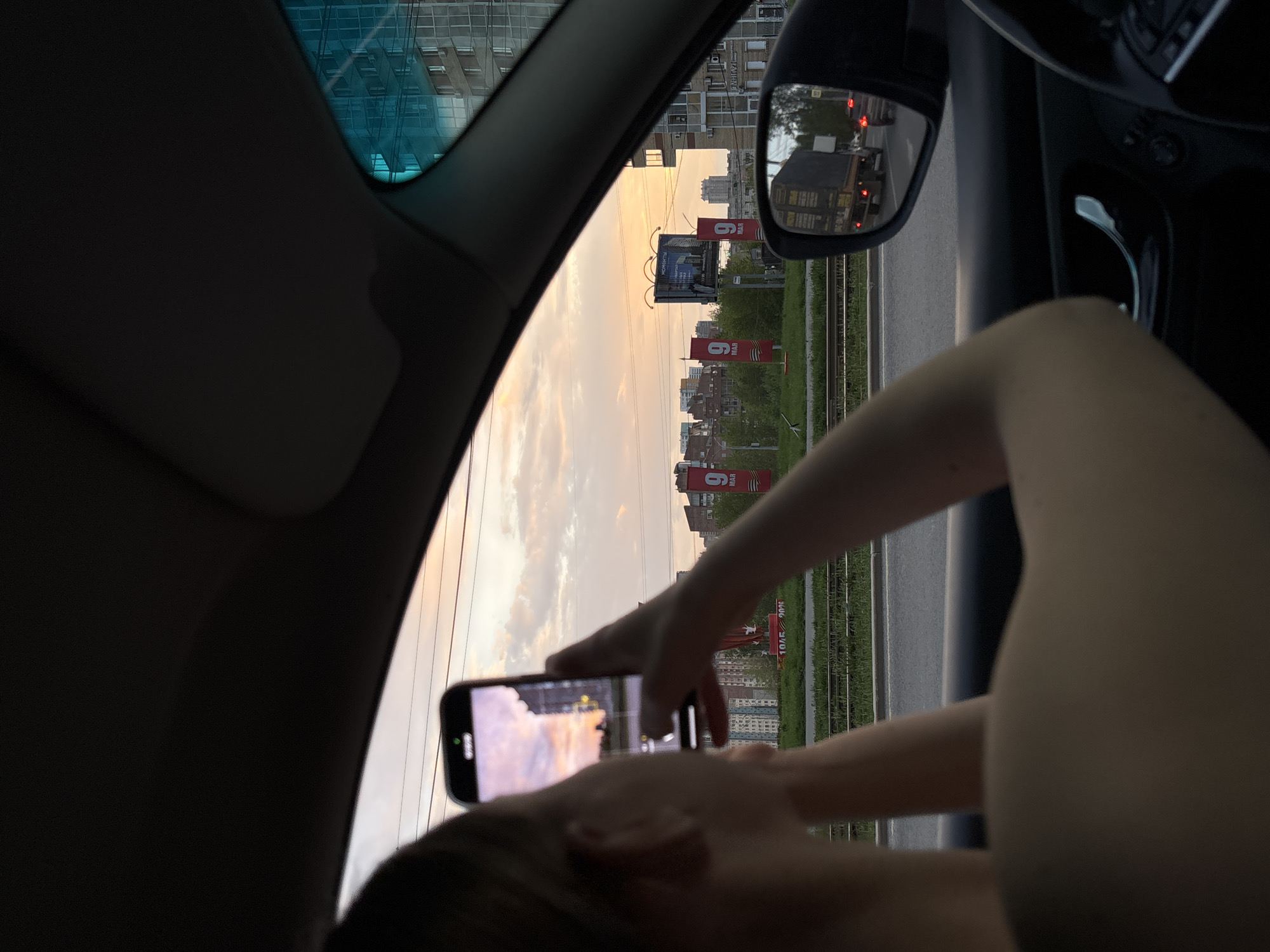 A person is holding a smartphone and taking a photograph of the cityscape outside of a car window. The skyline is marked by buildings, billboards, and red banners with the number "9" displayed. The side view mirror of the car is visible and reflects traffic. The picture is taken in Perm, Russia, evidenced by the banners containing the number 9. The person is likely a passenger in the car.