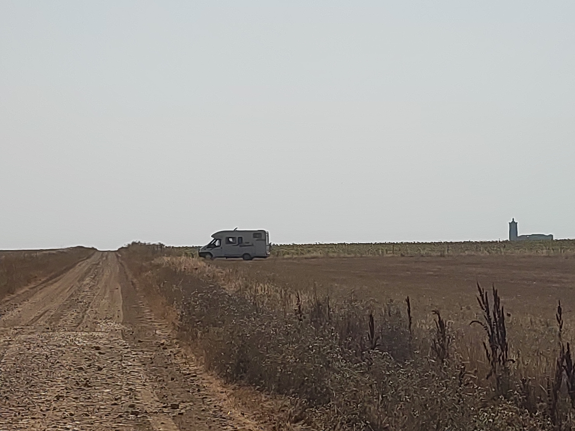 The image shows a rural landscape in Autillo de Campos, Spain. A gravel road stretches into the distance, flanked by dry vegetation. On the right side of the road, a gray camper van is parked in a field. Further in the distance, a tall, concrete structure with a dome-like top stands against a pale sky. The sky is overcast and hazy, suggesting a bright but muted day. The overall scene depicts a remote and quiet setting.