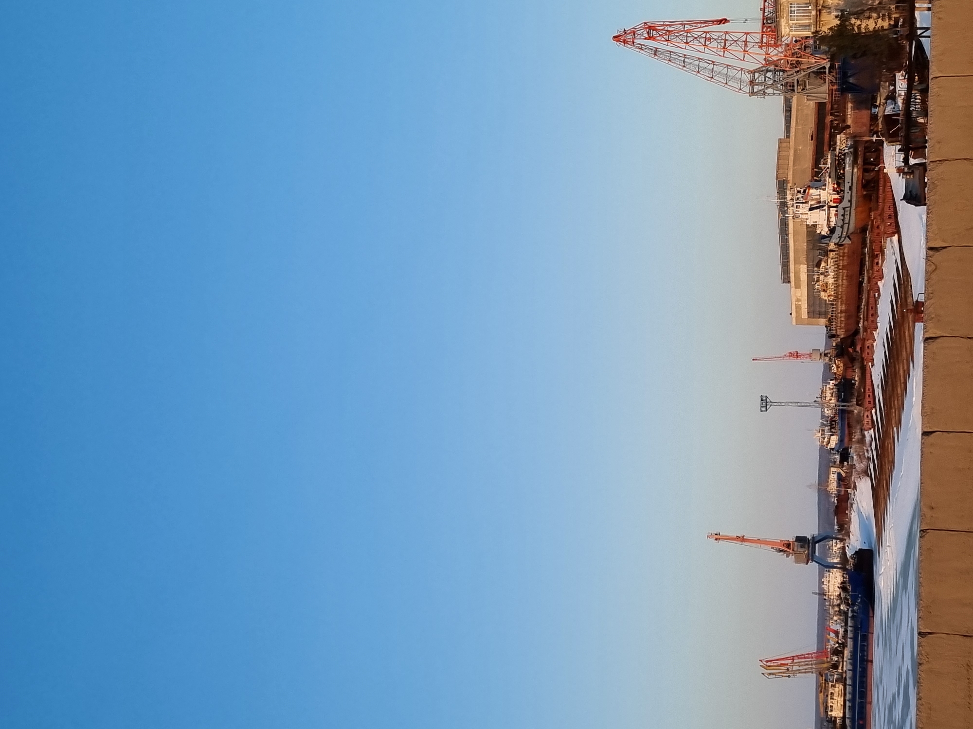 The image depicts a harbor scene in Chaikovsky, Russia. Several harbor cranes are visible, some orange and others red. There are also ships docked along a concrete pier. The sky above is clear and blue.