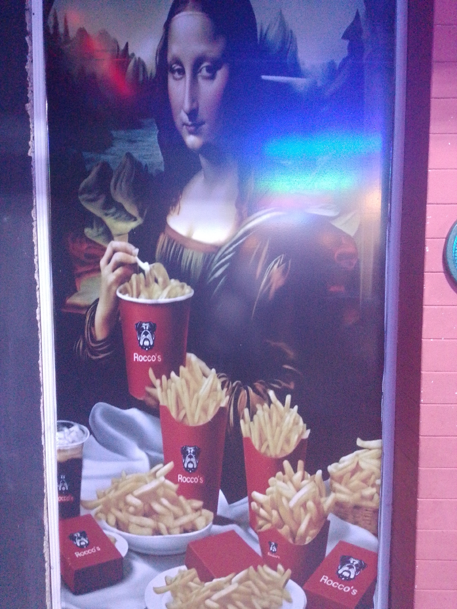 The image is a photograph of a poster depicting the Mona Lisa eating french fries. The fries are in containers and boxes labeled "Rocco's." The poster appears to be hanging on a wall, next to a pink tiled wall. The location is Tinaquillo, Venezuela, based on the poster’s presumed origin or establishment.