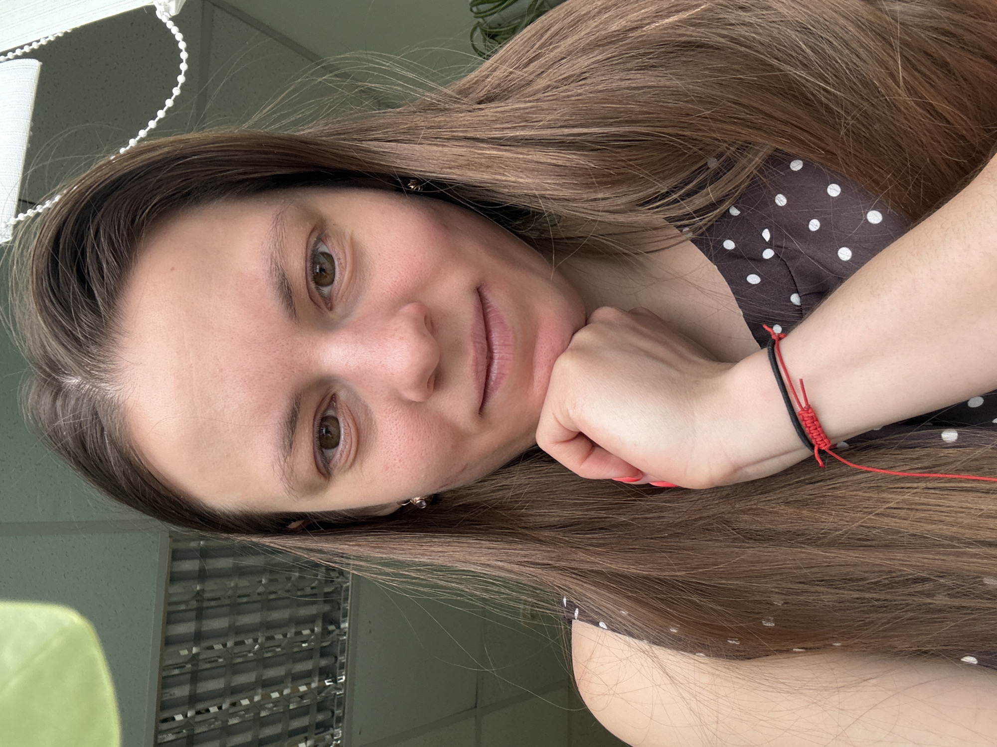 A woman with long brown hair is resting her chin on her hand. She is wearing a brown and white polka-dot dress and a red and black bracelet. The background appears to be an office setting, possibly in the city of Chaikovsky, Russia. A window with a blind can be seen.