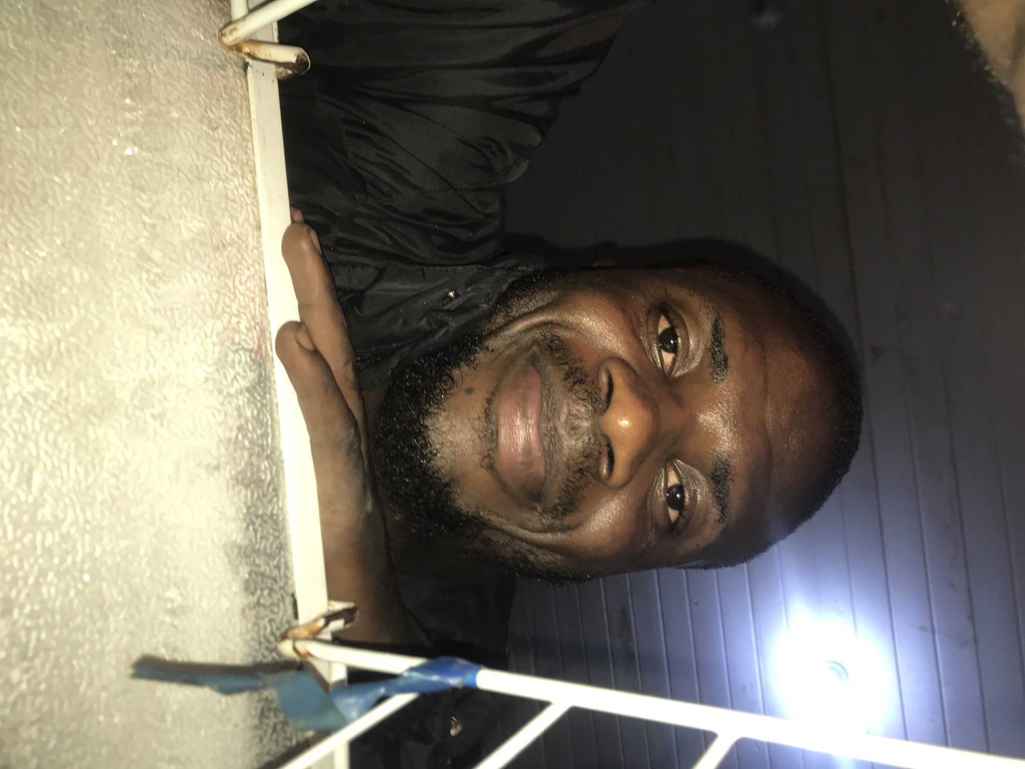 The image shows a close-up, low-angle shot of a Black man. He is wearing a black jacket and has a beard and short hair. He is smiling and looking directly at the camera. His left arm is bent and his hand is resting on a textured, light-colored surface. Behind him, the ceiling is visible, made of horizontal planks, and a bright light is illuminating the scene from the upper right. The image appears to be taken indoors, possibly at night, given the lighting conditions. The man's placement within New Stadium, Nigeria, is not specified by the visual information in the image.
