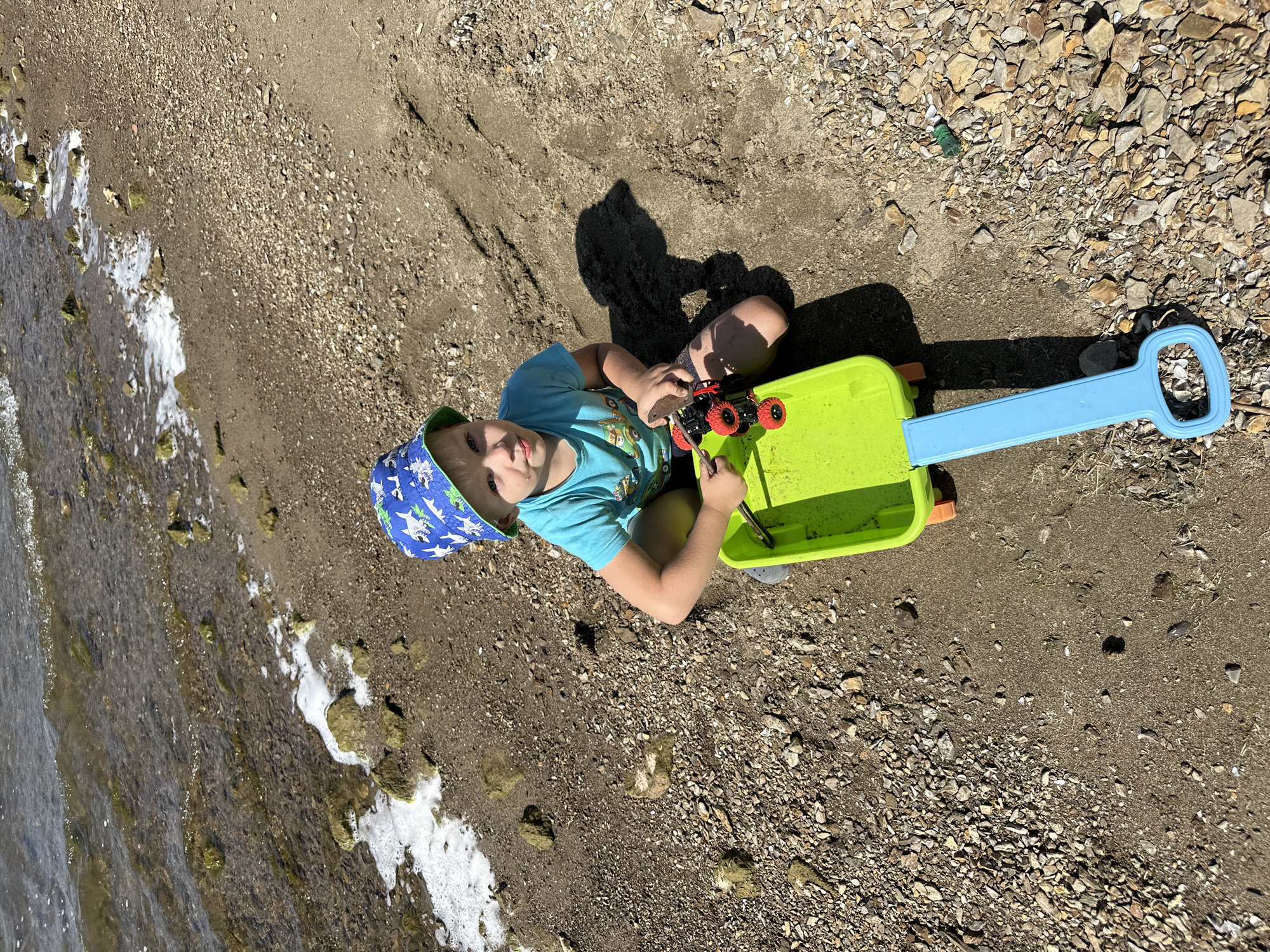In Gornyi Erik, Russia, a young child wearing a blue bucket hat with a shark pattern and a turquoise t-shirt kneels on a pebbly shoreline. The child is playing with a small, red toy car placed on a bright green wagon with a blue handle. The child's left hand is near the toy car and wagon, while their right hand is on the wagon. The environment consists of the water's edge with some foam and rocks, leading to a sandy and pebbly beach. The sun casts a shadow to the right of the child.