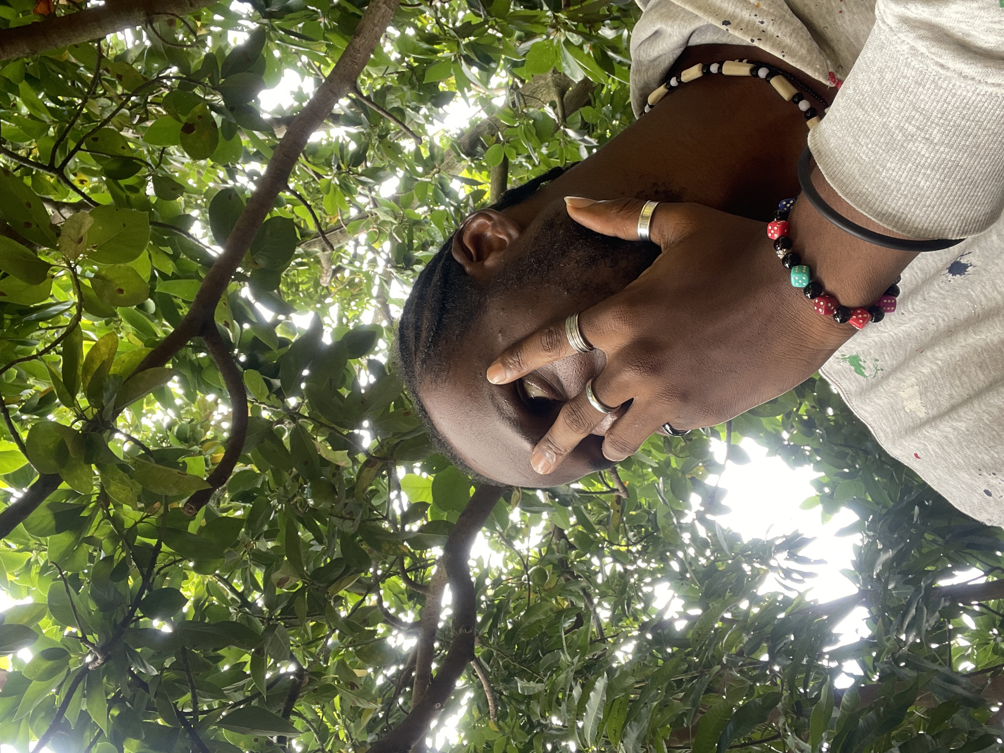 A close-up shot captures a Black person from the chest up, partially obscured by their right hand covering their face, in front of a backdrop of lush green leaves and branches. The person is wearing a light-colored hoodie with visible paint splatters, a beaded necklace, and a bracelet adorned with colorful dice and black beads. Two silver rings are visible on the hand covering the face. The setting appears to be outdoors, possibly in Jos, Nigeria, amidst dense foliage. The lighting suggests an overcast day, with sunlight filtering through the leaves. The image has a shallow depth of field, with the foreground sharply in focus and the background softly blurred.