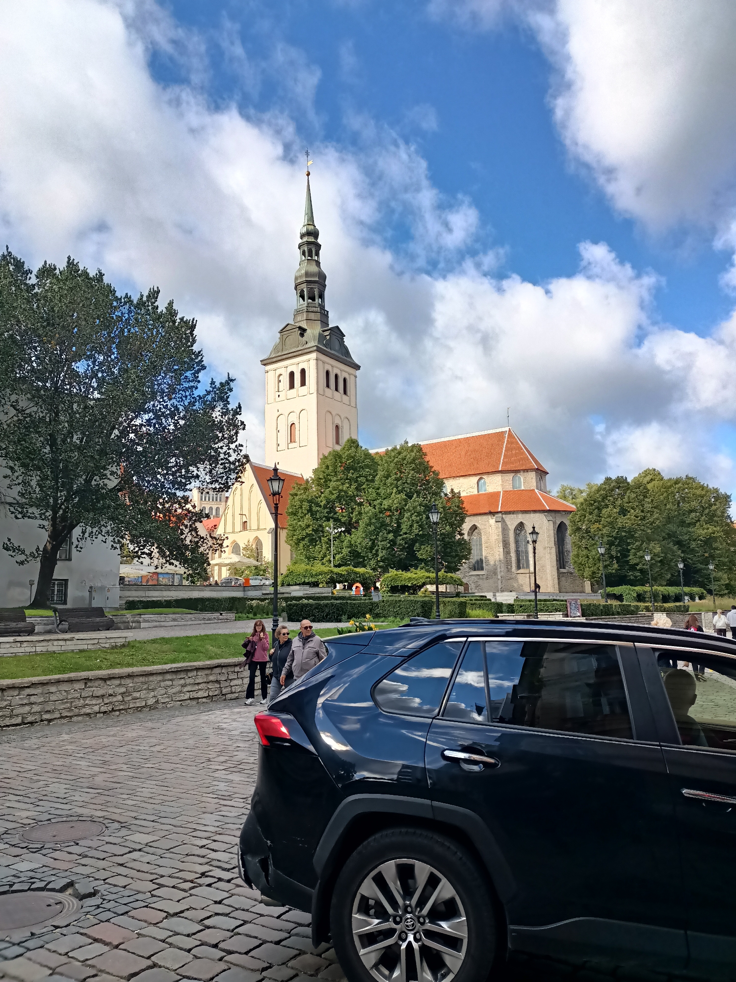The image shows a view of the St. Olaf's Church (Oleviste kirik) in Tallinn, Estonia. A black SUV is parked on a cobblestone street in the foreground, obscuring the lower portion of the church. The church features a tall, slender spire and a red-tiled roof. Trees and a stone wall with benches are visible in the middle ground. Several people are walking near the church. The sky is partly cloudy with blue patches.
