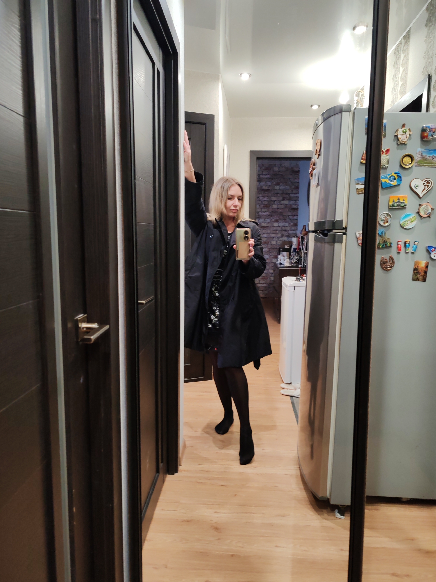 A woman stands in a hallway, likely inside an apartment, taking a selfie in a mirror. She is wearing a black coat over a dress, with black tights and shoes. Her left arm is raised near the door frame, while she holds a phone in her right hand. The background includes a refrigerator covered in magnets, a doorway leading to another room with exposed brick walls, and several doors, one of which is slightly ajar. The floor is wooden. The location appears to be an urban domestic setting, but there's no clear evidence to confirm the specific city of Petropavlovsk-Kamchatsky, Russia.