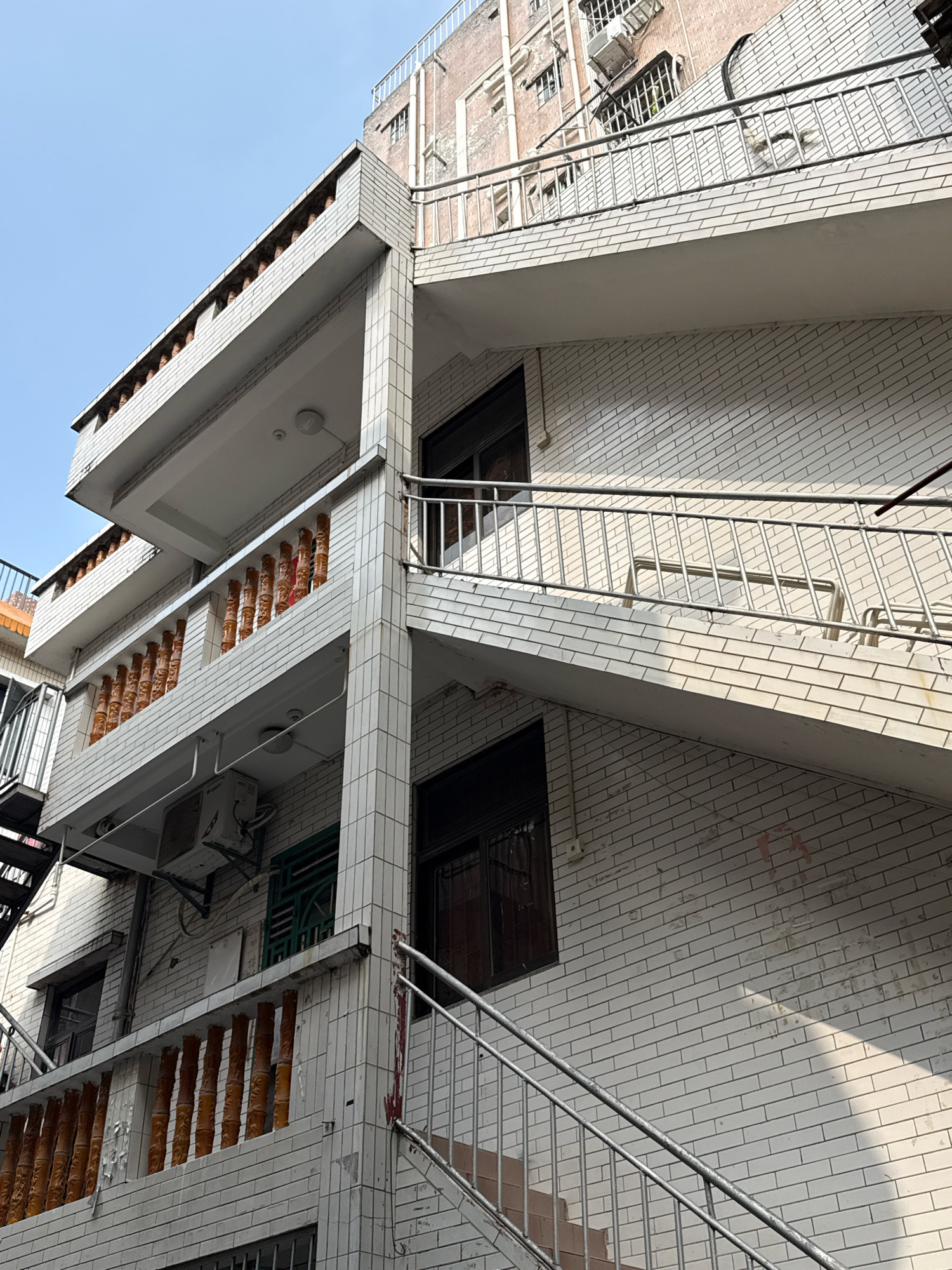 The image depicts the exterior of a multi-story building, likely an apartment complex, in Nanshan District, China. The building is constructed with white brick and features several balconies with decorative railings. An external staircase, made of metal, ascends along the side of the building. Windows with dark frames are visible on different levels, and an air conditioning unit is mounted on one of the walls. The sky above is clear and blue. The overall scene conveys a residential environment with typical urban architectural elements.
