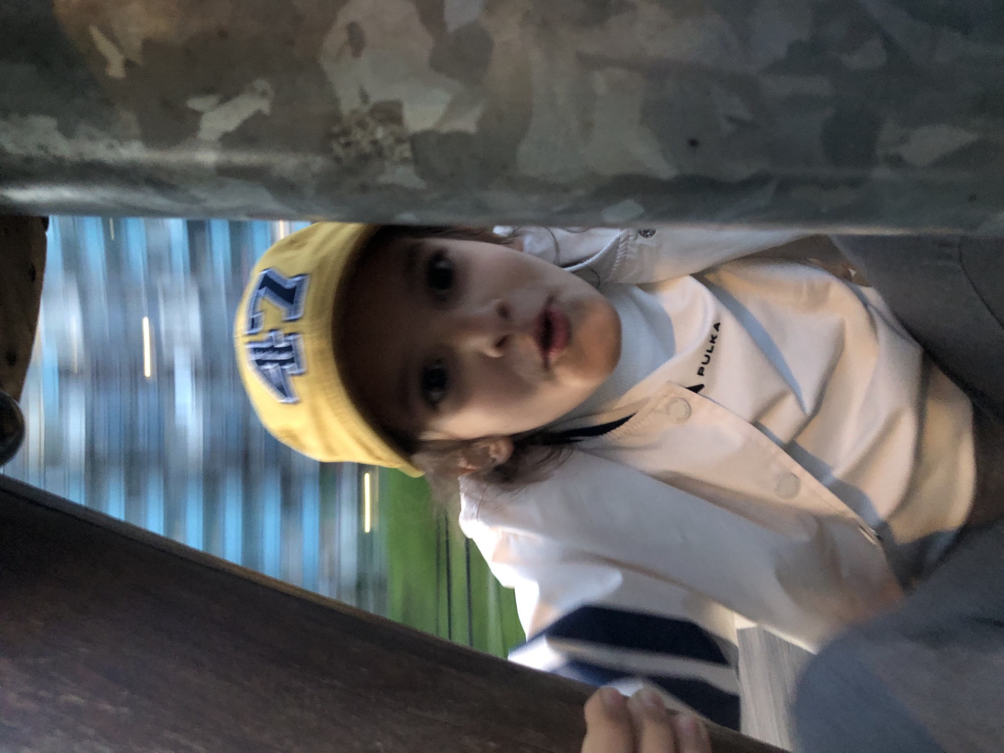 A young child with dark hair and eyes is captured in a close-up, low-angle shot, peeking directly at the camera. The child's head is tilted significantly to her left, or the camera was held at an angle. She wears a bright yellow knit beanie adorned with dark, stylized letters, possibly a logo. Over a white long-sleeved top, she wears a white lightweight jacket featuring subtle light blue vertical stripes or seams, with the brand name "PULKA" printed in black text near her right shoulder. Her mouth is slightly open, conveying an inquisitive or curious expression.
The child appears to be looking out from a gap between two dark, blurry, out-of-focus horizontal structures that frame the top and bottom of the image, suggesting she might be inside a playground tunnel, slide, or beneath a railing. In the background, a soft blur of vertical blue and white stripes or elements is visible, possibly belonging to a building or a distant structure, interspersed with hints of green, suggesting an outdoor setting. The lighting is soft and even, indicating possibly an overcast day or late afternoon. The scene evokes a candid moment of childhood play or exploration in an urban or park environment, consistent with a location like Saint Petersburg.