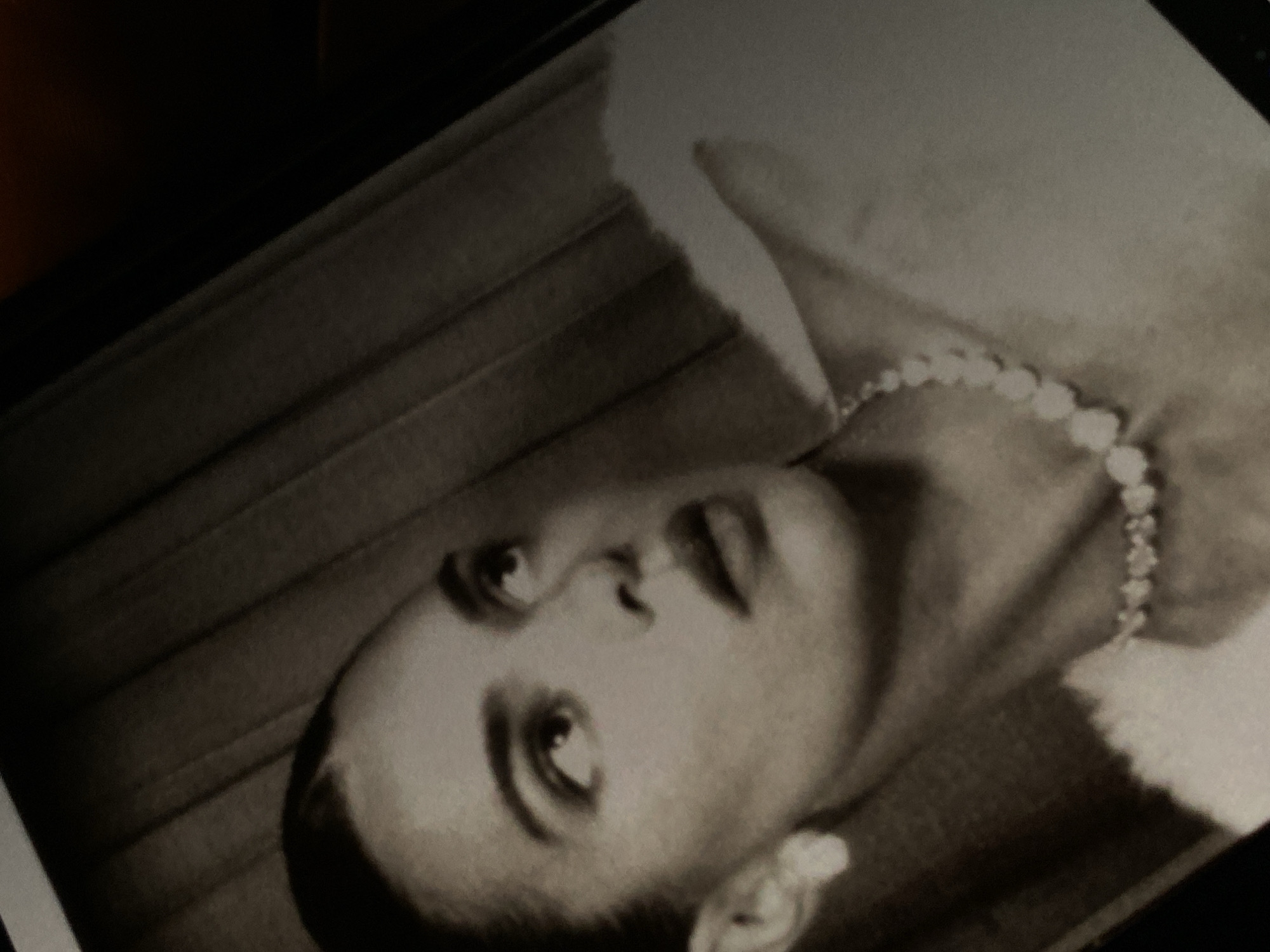 A black and white photograph depicts a person with short, dark hair wearing a pearl necklace. The person appears to be wearing a fur collar or wrap. The background is blurred, and may show drapery. The location is unknown.