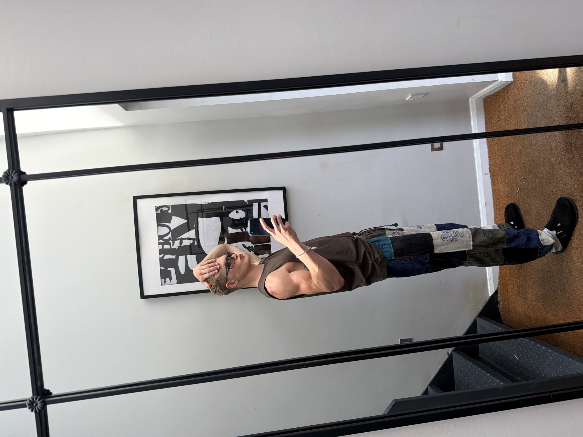 A reflection in a large, black-framed mirror shows a fair-skinned man taking a selfie with a smartphone. He is wearing sunglasses, a brown tank top, and patterned pants. The background includes a white wall with a black-framed piece of abstract art, a portion of brown carpet, and a set of black metal stairs. The reflection reveals a room with minimalist decor. The location cannot be determined with the given information.