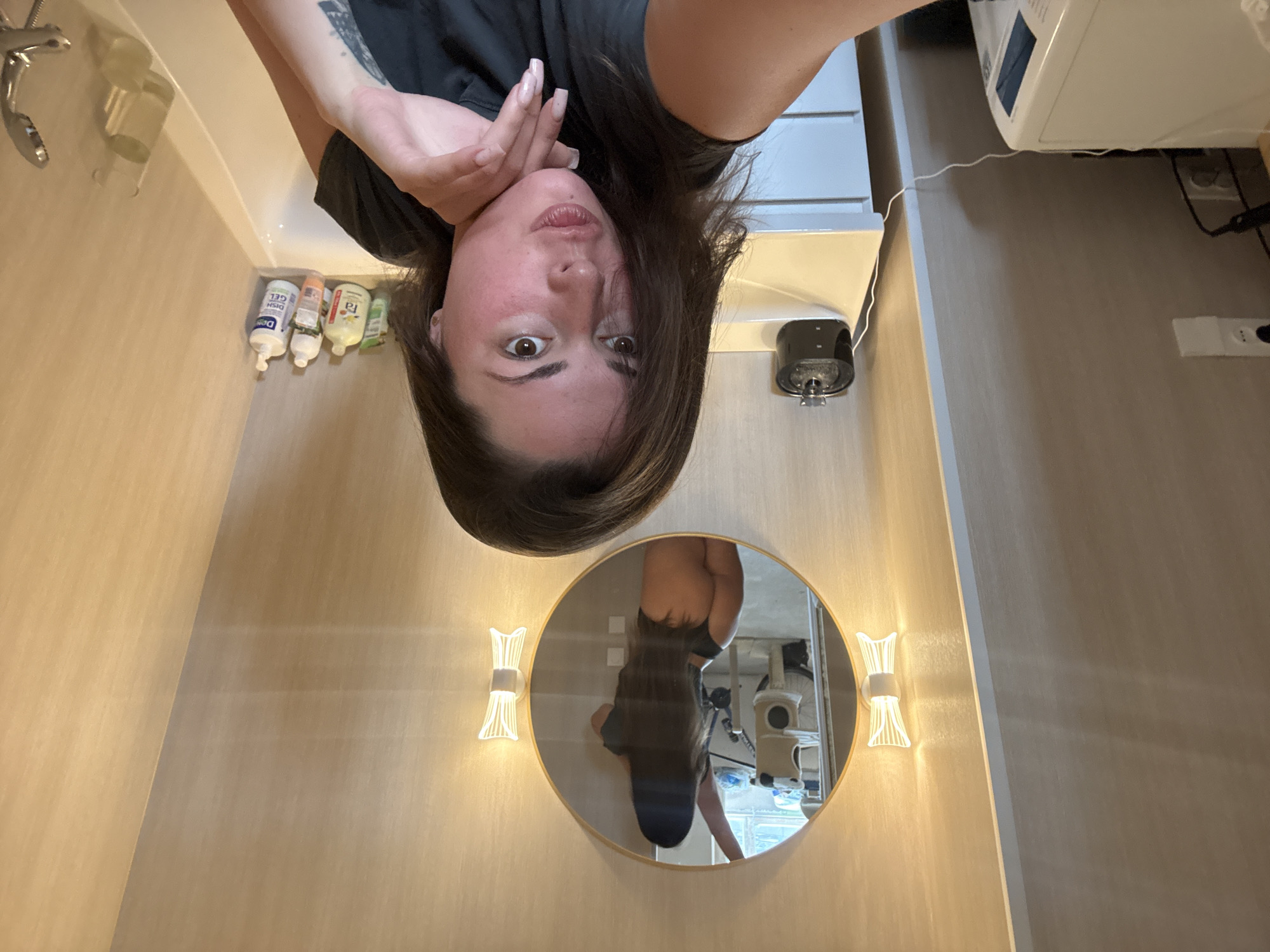 The image shows a woman taking a selfie in a bathroom. The woman is wearing a black t-shirt and has dark brown hair. She is leaning on her hand and looking at the camera. In the background, there is a round mirror reflecting another woman, who is wearing a black top and black underwear and posing with her back to the mirror. The bathroom has light-colored walls and a white sink. There are two wall-mounted lights on either side of the mirror. On the counter next to the sink, there is a black device and several bottles of toiletries. To the left of the sink, there is a white washing machine.