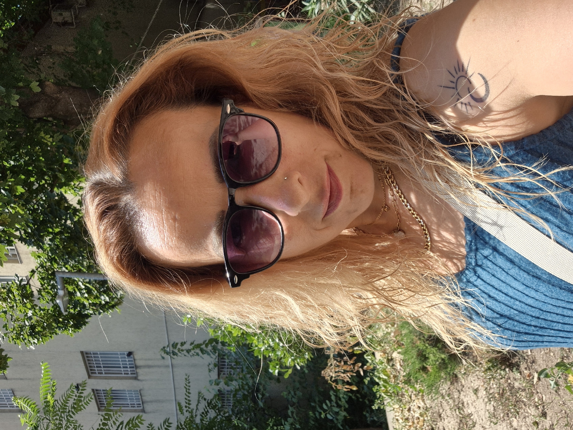 A medium shot captures a woman with strawberry blonde, wavy hair, wearing sunglasses and a blue top. She has a nose ring and a tattoo of a sun and crescent moon on her upper left arm. She is outdoors, with a building and trees in the background. The lighting suggests it is daytime.