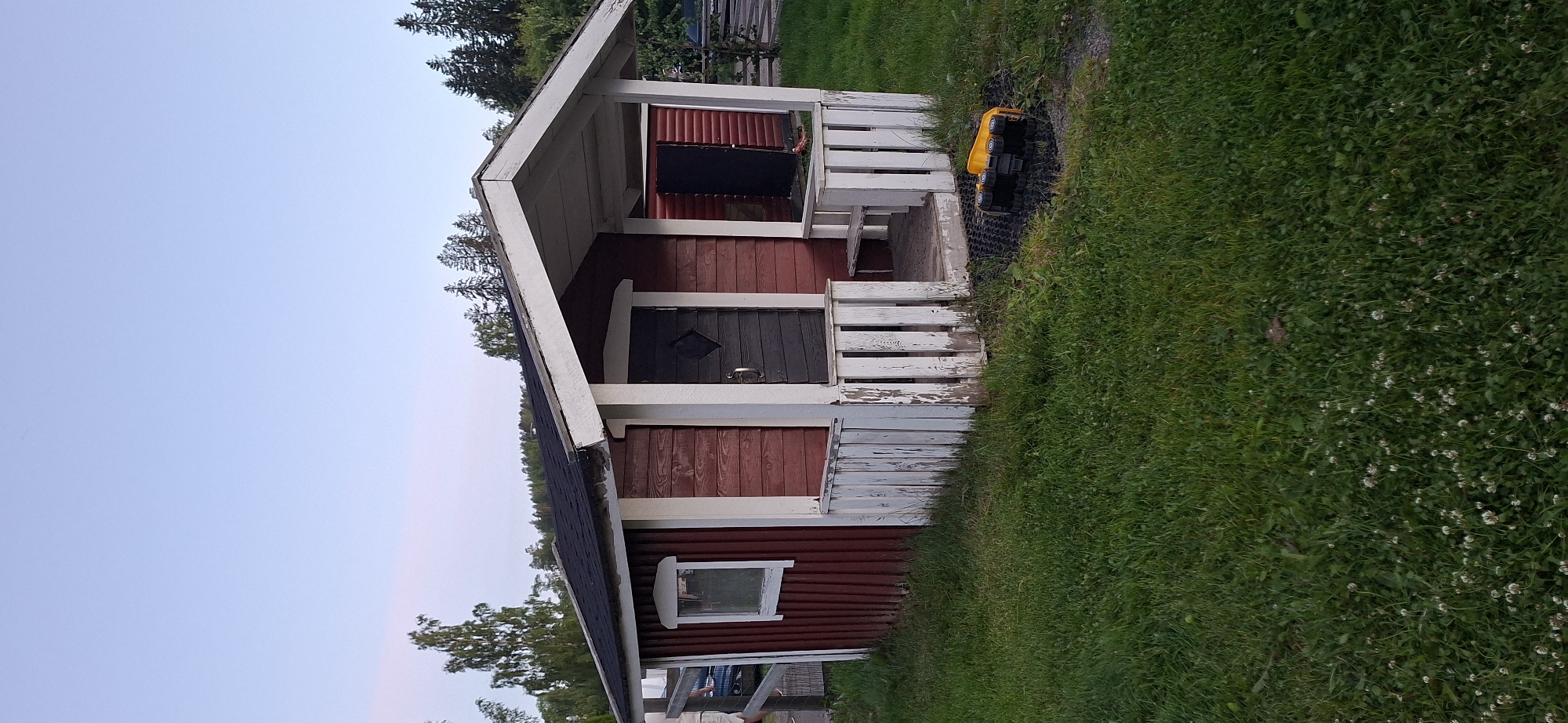 The image captures a small, weathered red wooden cabin with a dark-colored door adorned with a diamond-shaped detail and a silver handle. The cabin features a white-trimmed window on its left side. A white, peeling wooden fence encloses a small porch area leading to the cabin's entrance, which includes a wooden step and a built-in bench. To the right of the porch, another dark-colored door is visible. The cabin sits on a patch of green grass, with a small yellow toy truck overturned on a black mat in front of the porch. In the background, trees line a hill, and the sky is a pale blue, suggesting it is either early morning or late evening. The overall setting appears to be a rural or suburban environment, possibly a backyard or a small park area within or near the city of Salpakangas, Finland, given the presence of coniferous trees and deciduous trees typical of the region.