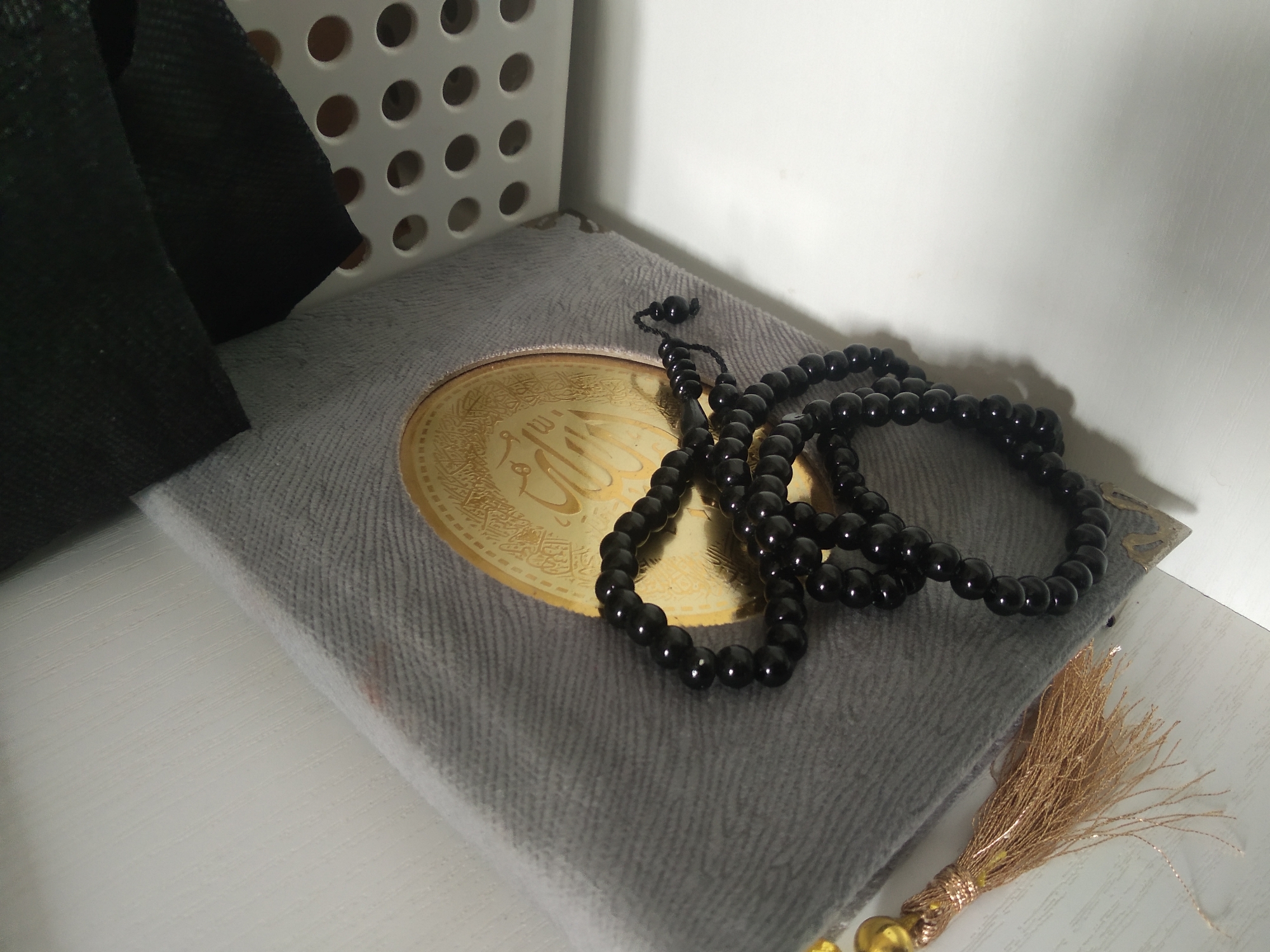 The image shows a close-up view of Islamic prayer beads made of black stones, placed on top of a gray book. The book has a circular gold inlay with Arabic calligraphy. To the left of the book, a black fabric object is visible, and behind it, a white plastic container with circular holes. A gold tassel hangs from the bottom right corner of the frame. The scene appears to be on a white, textured surface, possibly a shelf or table, with a white wall in the background. The lighting suggests an indoor setting with light coming from the right, casting shadows. There are no people present in the image. The overall impression is one of religious items arranged for display or use. The location is not identifiable from the visual information provided.