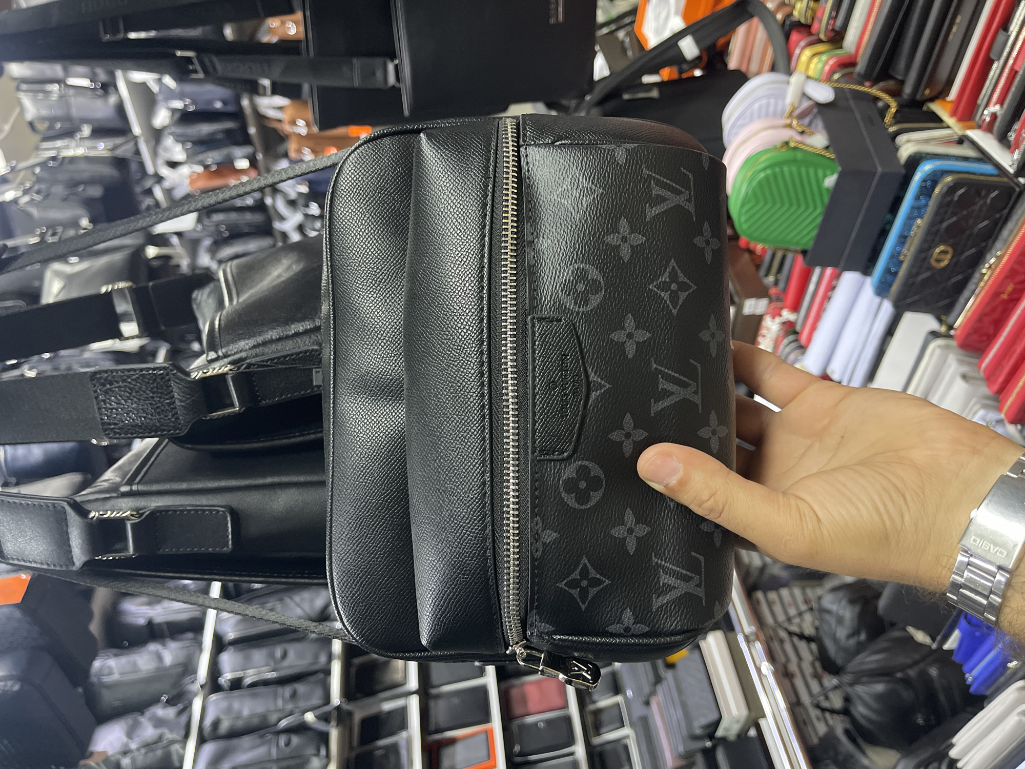 The image shows a person's hand holding a black patterned crossbody bag in what appears to be a market stall. The bag has a prominent "LV" monogram pattern and a silver zipper. In the background, shelves are filled with various bags and accessories, including smaller colorful clutch bags in a box. The overall environment suggests a retail setting where bags and accessories are being sold. The prompt mentions Moscow, Russia, but there are no visual cues in the image to confirm this specific location.