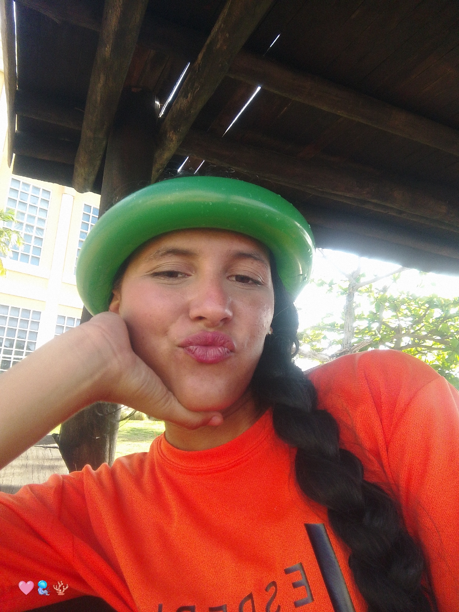 In a selfie taken outdoors in Pedro González, Venezuela, a woman is pictured from the chest up. She is wearing an orange t-shirt and a green inflatable hat. Her right hand is resting on her chin and she is puckering her lips. A black braid of hair extends down her right side. In the background, a wooden structure can be seen overhead, along with a building with white-paned windows and foliage. The lighting suggests it is daytime.