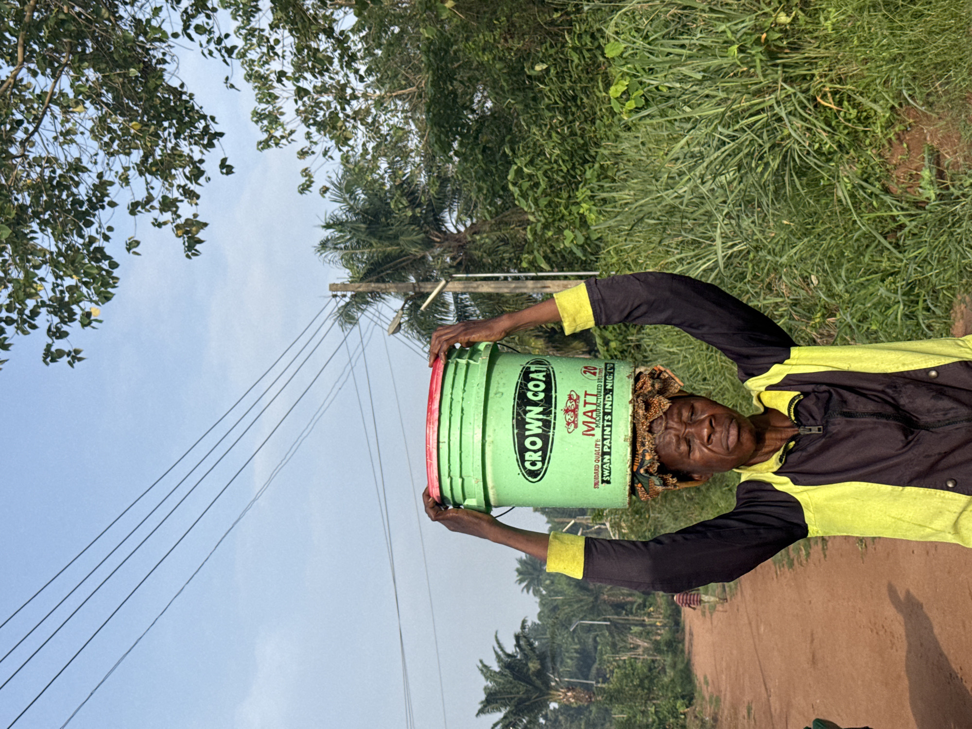 An outdoor, eye-level shot captures a person carrying a green bucket on their head along a dirt path in Eziudo, Nigeria. The person, who appears to be an older adult with dark skin, wears a black and yellow long-sleeved shirt and a patterned head wrap beneath the bucket. Their face is turned towards the camera, showing a neutral expression. Their left arm is raised, with fingers touching the side of the bucket, possibly to steady it.
The green bucket has a red rim and features prominent text, including "CROWN COAT," "MATT 20," "STANDARD QUALITY," and "SWAN PAINTS IND. NIG. LTD."
The setting is rural or semi-rural, characterized by a wide, light brown dirt road that recedes into the distance, where another figure can be faintly seen. Lush green vegetation, including tall grass and various trees (some leafy, some resembling palm trees), frames the path on both sides and in the background. Overhead, power lines extend across a clear, pale blue sky with minimal cloud cover, suggesting daytime. A utility pole is visible to the left of the main subject. The overall impression is one of daily activity in a vibrant natural environment.
