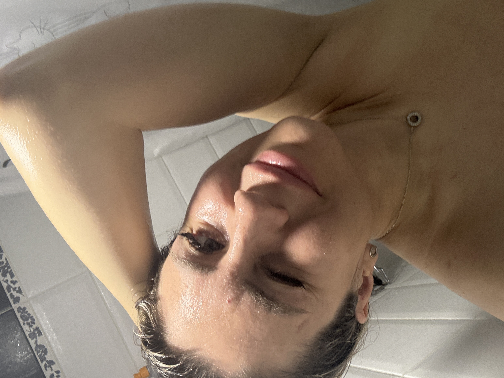 A woman is taking a selfie in a shower. She is looking at the camera with her head tilted and her arm behind her head. The shower is tiled with white tiles and has a decorative border. The woman has wet hair and is wearing a necklace. There is a bottle of shampoo visible in the background. There is no information available about the specific location within Novoaltaysk, Russia.