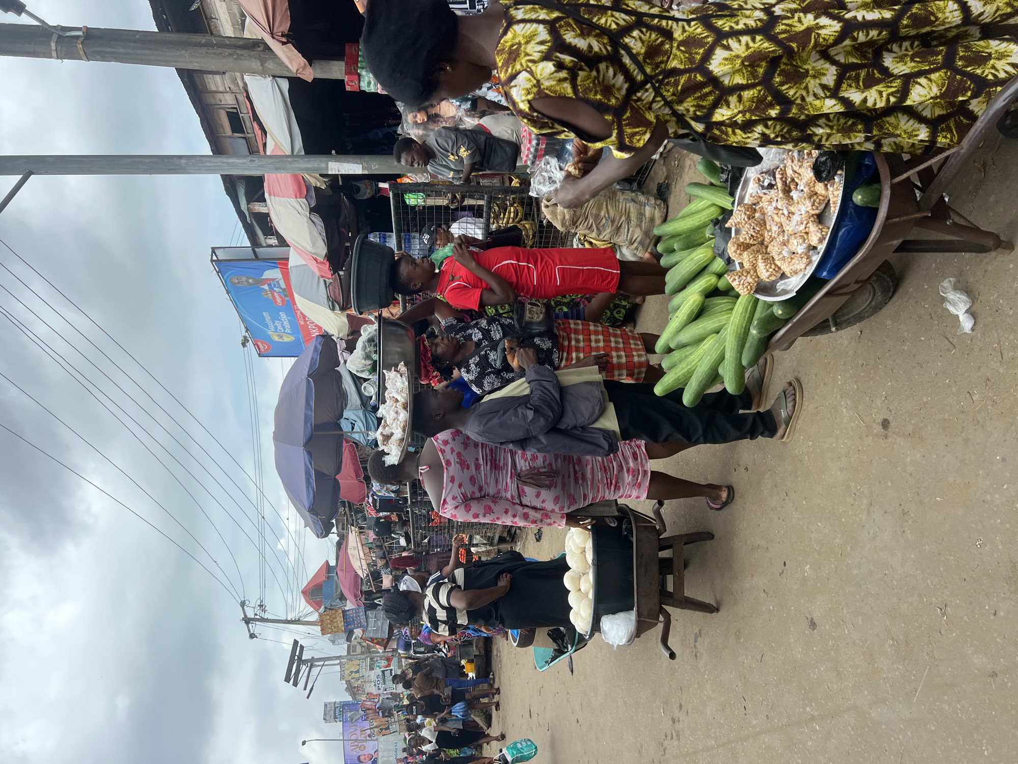 The image depicts a bustling outdoor market scene in Nkpor, Nigeria, under a cloudy sky. Several individuals are engaged in commerce, with a particular focus on vendors selling food items. In the foreground, a woman in a yellow and brown patterned dress stands near a wheelbarrow filled with cucumbers and a platter of what appears to be fried snacks, possibly peanuts. Next to her, a young boy in a grey jacket and black pants stands by the wheelbarrow. Behind him, a woman in a pink floral dress and a striped-shirted individual stand near a wheelbarrow containing round, white food items, likely dumplings or dough. Other vendors and shoppers are visible in the background, amidst stalls and awnings, creating a dynamic marketplace atmosphere. Overhead, electrical wires crisscross the sky, a common sight in urban Nigerian environments. The ground is a dusty, unpaved surface.