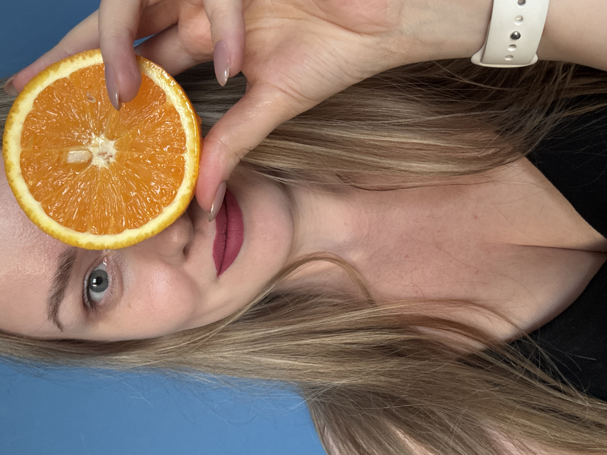 A fair-skinned woman with light brown hair is holding a slice of orange over her left eye. The woman is wearing mauve lipstick. Her left wrist has a white Apple Watch band. The background is blue. The location of Chaikovsky, Russia is not depicted or inferable from the image.