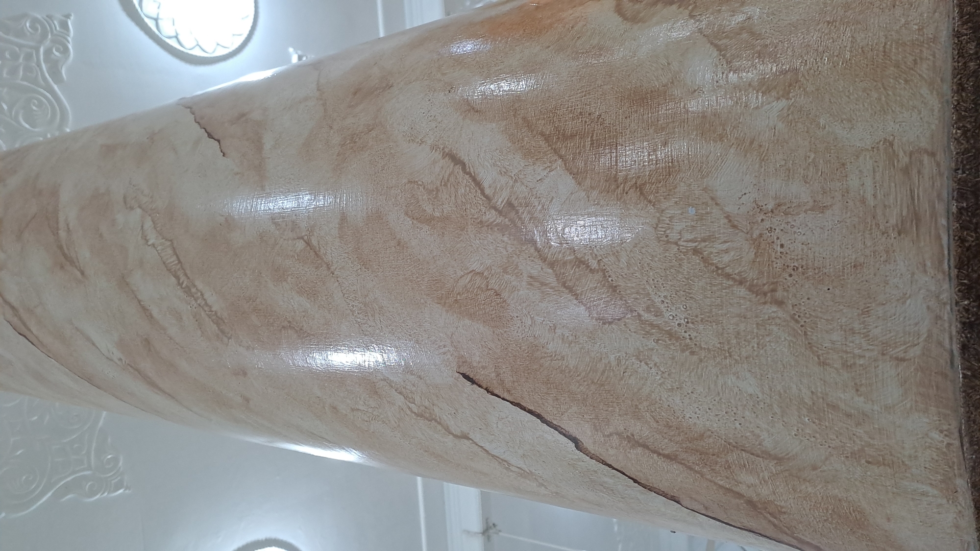 The image shows a close-up of a textured column with a marble-like finish. The column is curved and appears to be part of a larger structure, possibly an interior space. The texture is characterized by swirling patterns of light brown and beige, with some darker brown accents that resemble natural stone veins. The surface is glossy, reflecting light in irregular patches.
In the upper left and lower left of the image, there are glimpses of white architectural elements, including what appear to be decorative moldings and an oval light fixture. These elements suggest an indoor setting with some form of ornate design. The lighting appears to be coming from the fixtures, creating highlights and shadows on the column's surface, indicating it might be daytime or a well-lit interior.
The texture on the column shows a variety of brush strokes or application techniques, creating a dynamic and uneven surface. There are areas with fine, linear textures and others with a smoother, blended appearance. A thin, dark crack is visible on the surface of the column, running diagonally across a section of the image. The right edge of the image shows a darker, rougher texture, possibly a carpet or a different material adjacent to the column.
There is no visible text in the image. The focus is entirely on the architectural detail and the tactile quality of the column's finish. The overall impression is one of a detailed study of an interior architectural element.