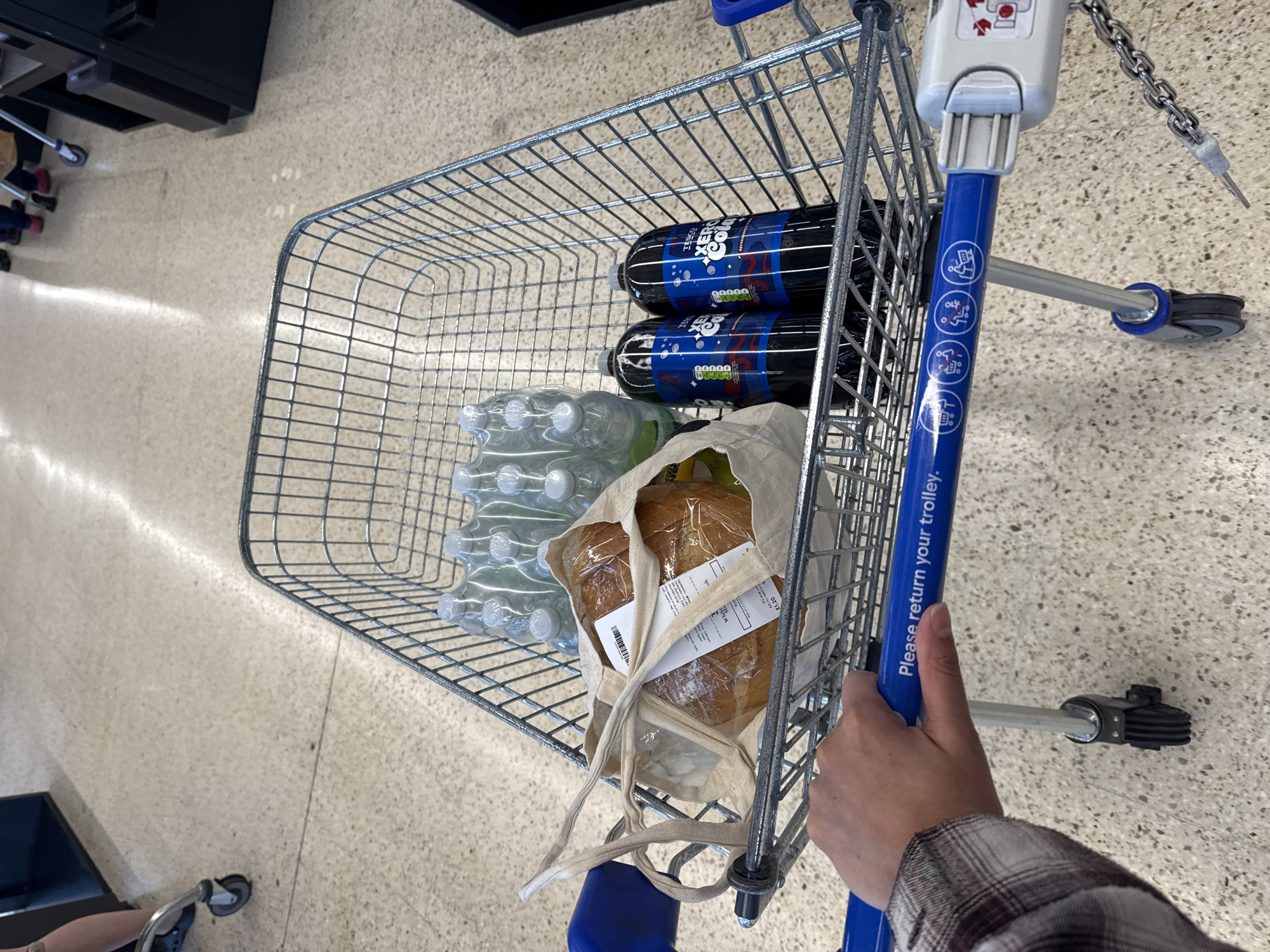 A first-person view photograph shows a shopping trolley filled with groceries. Inside the trolley are two large bottles of Tesco Zero Cola, a multipack of water bottles, and a loaf of bread in a reusable canvas bag. The trolley has a blue handle and a chain attached, indicating it's likely from a supermarket. A person's hand, wearing a plaid sleeve, grips the trolley handle. The floor is a light-colored, speckled tile, suggesting an interior space, possibly a supermarket in Great Yarmouth, United Kingdom.