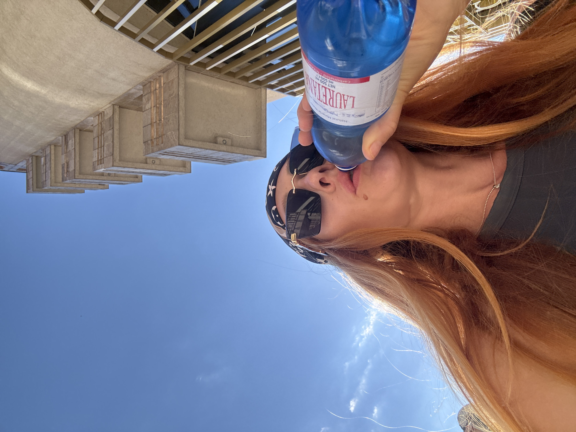 The image shows a person drinking from a blue bottle of Lauretana natural mineral water. The bottle is held up to their mouth, and the person is wearing sunglasses and a bandana. Their long, reddish-blonde hair is flowing. The background consists of a clear blue sky with some wispy clouds, suggesting it is daytime with good weather. In the upper portion of the image, there is a glimpse of what appears to be a concrete structure with some wooden beams, possibly part of a building or a modern architectural element. The overall scene evokes a sense of relaxation and refreshment, possibly in an outdoor setting given the bright sky. The bottle has text on its label, including "LAURETANA," "NET 500 ml," and "Natural Mineral Water." The image is oriented sideways.