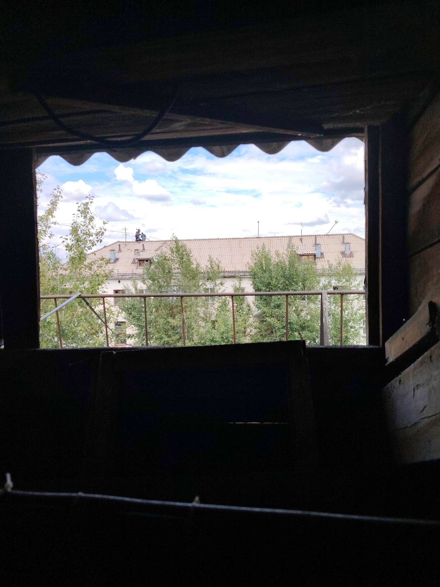 This image shows a view from within a dark, enclosed space, looking out through a rectangular opening at a partly cloudy sky, a building, trees, and a metal railing. The opening has a wavy, corrugated edge at the top. In the background, a light-colored building with a tiled roof is visible. Two people are seen on the roof of this building, appearing to be working. Green trees are in front of the building, and a rusty metal railing runs horizontally across the middle of the view. The foreground is filled with dark, indistinct shapes, suggesting the interior of a structure. The image's details are consistent with an outdoor scene in Astana, Kazakhstan, possibly from a balcony, attic, or abandoned structure overlooking a residential area.