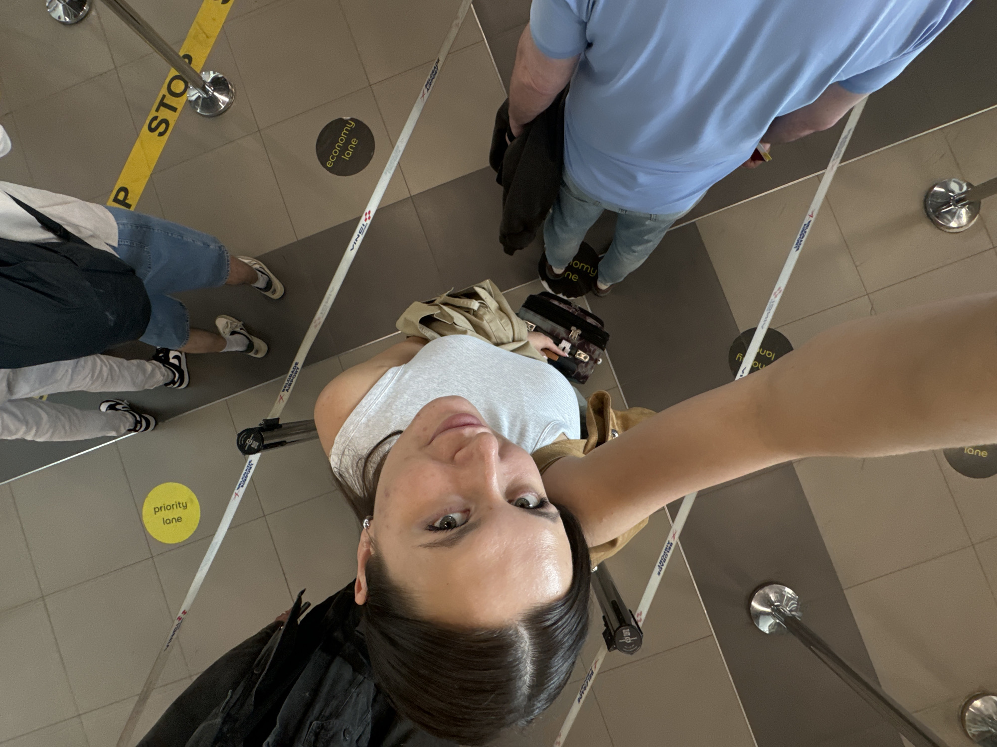 Here is a concise description of the image:
The image shows a selfie taken by a woman in what appears to be an airport queue. The camera is pointed upwards at an angle, capturing the woman's face and the surrounding environment. She is standing in a "priority lane" at Belgrade Airport in Surcin Urban Municipality, Serbia. Other individuals are also in the queue, with their legs and lower bodies visible. The queue is managed by stanchions and retractable belts, and there are floor markings indicating "economy lane" and the word "STOP". The floor is tiled in a neutral color.