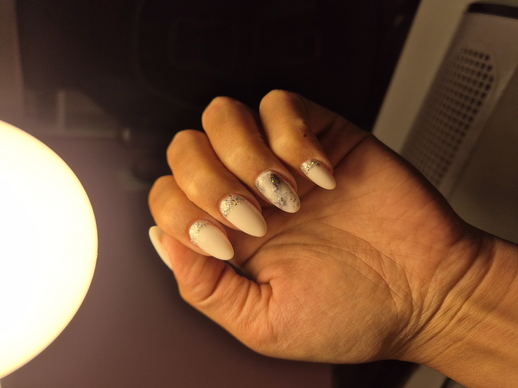 A close-up shot features a human hand, likely female, showcasing a freshly manicured set of nails. The hand is positioned centrally, with fingers slightly curled to present the nail art. The skin has a warm, tanned appearance, accentuated by a strong, circular, artificial light source, possibly a lamp, emanating from the left side of the frame.
The nails are almond-shaped and painted with a pale, creamy beige or off-white polish. The ring finger and middle finger display a glittering design, with silver or gold sparkles concentrated at the cuticle and diffusing towards the tips. The index finger features a distinct white and grey marbled or abstract pattern, subtly highlighted with gold flecks. The thumb and pinky nails are uniformly coated in the pale polish.
The background is dark and largely out of focus, suggesting an indoor environment, potentially at night or in low light. A faint, rectangular object with a discernible grid pattern, possibly an air purifier or similar appliance, can be seen in the blurred background on the right. No visible text is present in the image. The scene is situated in Redina, Bulgaria.