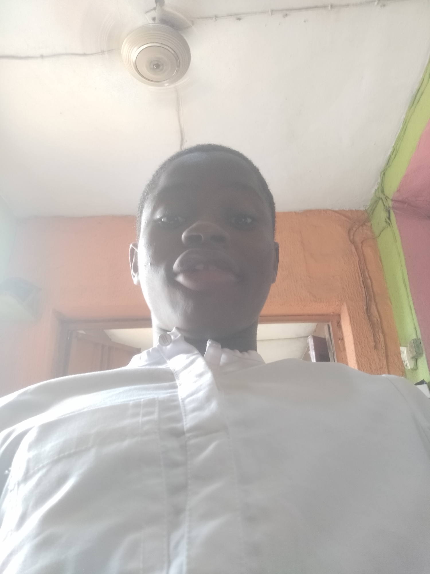 A close-up selfie taken from a low angle, featuring a young person with dark hair wearing a white, buttoned shirt. Above the person is a white ceiling with a ceiling fan. In the background, there is a doorway and walls painted in orange, green, and pink. The location is Unknown, Unknown.