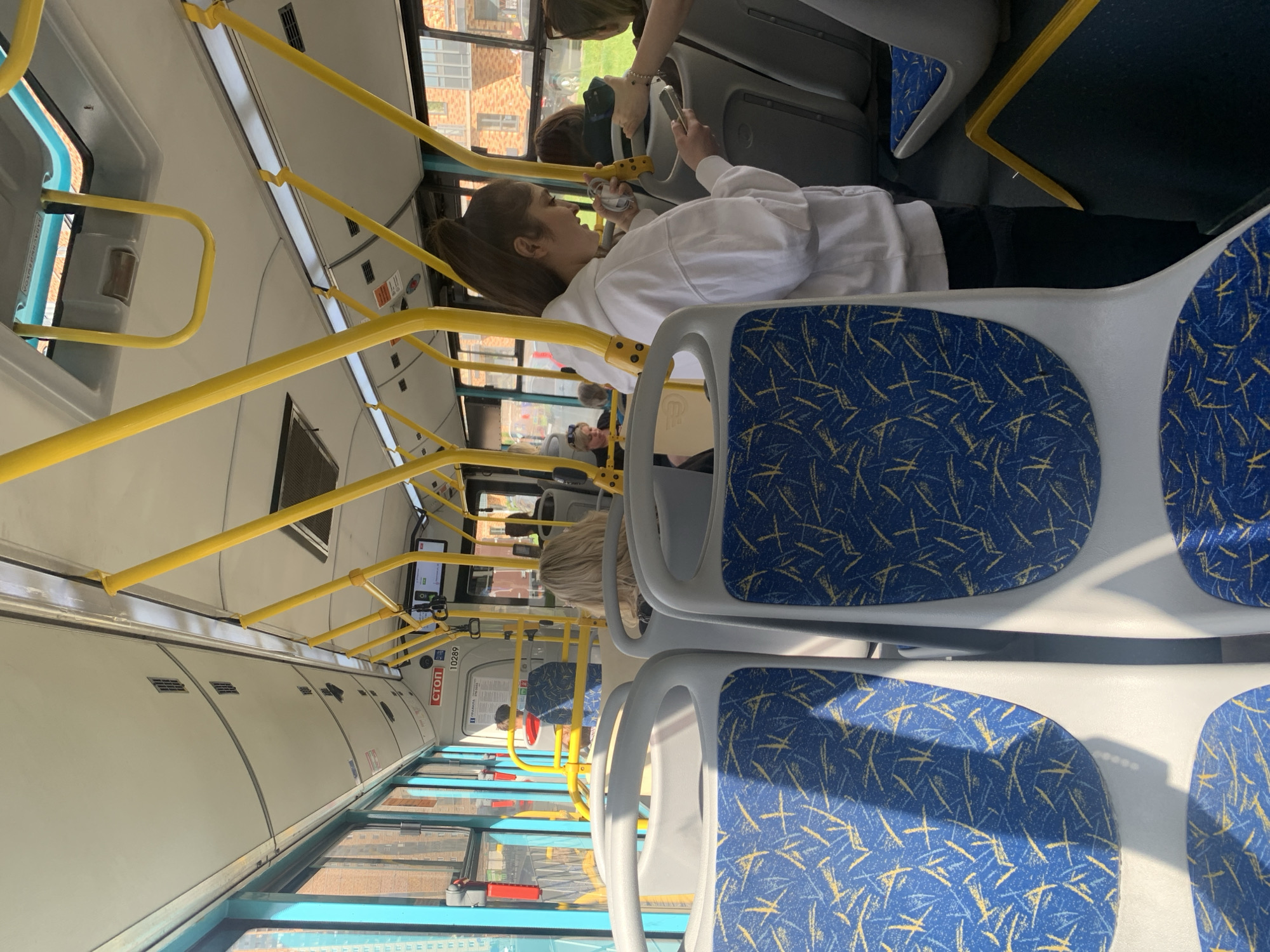 The image shows the interior of a bus, likely in the city of Ladozhskoe Ozero, Russia, given the presence of a sign with "СТОП" (Stop in Russian). The seats are upholstered in blue fabric with a yellow abstract pattern. Yellow metal poles are visible throughout the bus, serving as handrails. In the foreground, two seats with the blue and yellow patterned upholstery are prominent. A young woman in a white hoodie is standing in the aisle, looking at her phone and holding a white object, possibly earbuds. Other passengers are visible in the background, some seated and some standing. Sunlight streams in through the windows, illuminating the interior. The environment appears clean and well-maintained.