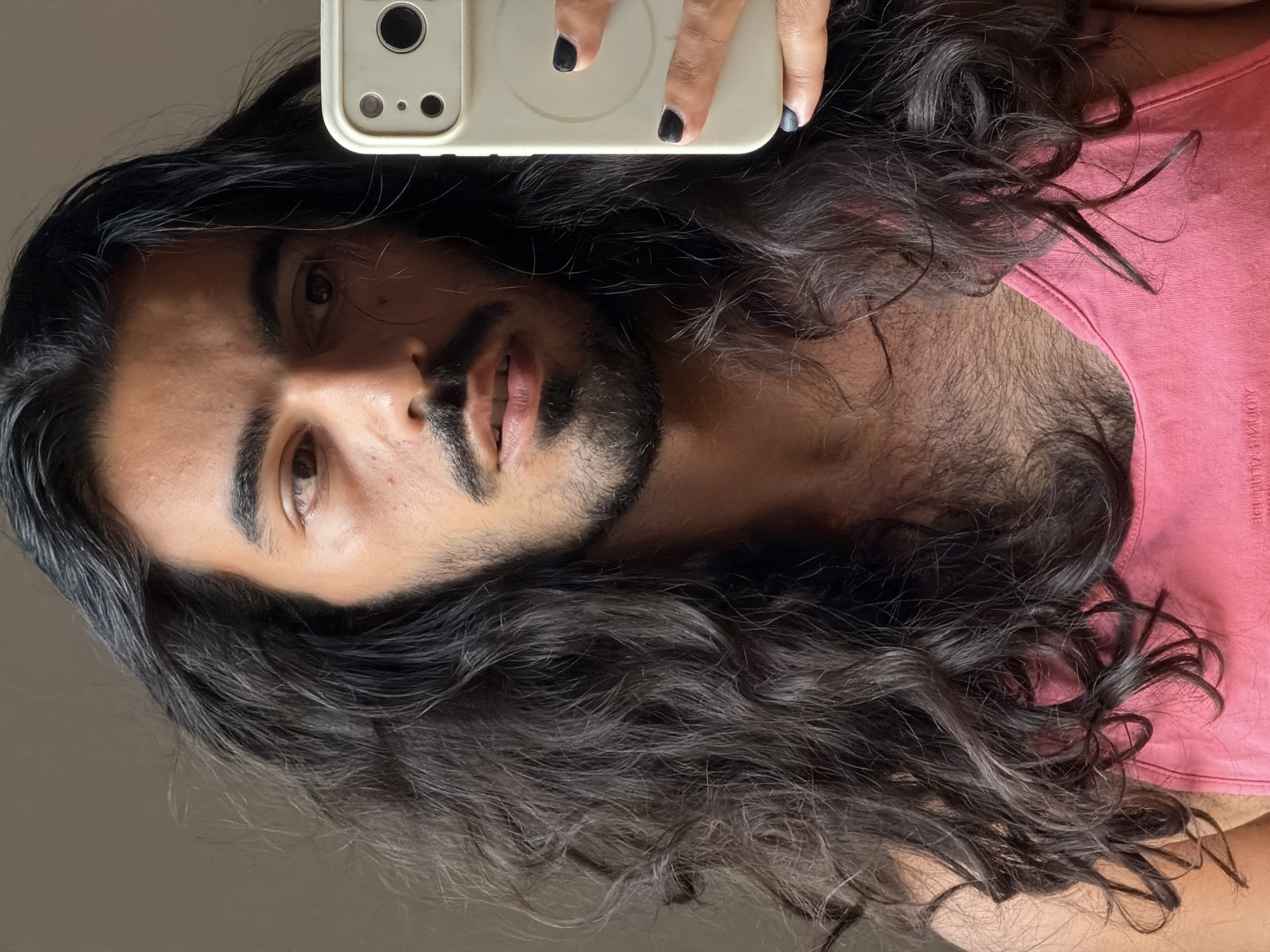 The media file is a close-up, vertically oriented selfie taken by a person with long, dark, wavy hair. The subject is looking towards the camera, with their face and chest visible. They are wearing a pink tank top. The subject's hands are also visible at the top of the frame, holding a smartphone with a light-colored case. The phone has a visible camera array. The background appears to be a plain, neutral-colored wall or surface, suggesting an indoor setting. The lighting is soft and even, indicating it may be daytime, possibly indoors with natural light. There is no clear indication of the weather or specific time of day beyond that. The overall mood conveyed by the image is casual and personal, typical of a selfie. The subject's expression is neutral. No text is visible in the image. Based on the provided location context of North Kuta, Indonesia, it is plausible this photo was taken there.