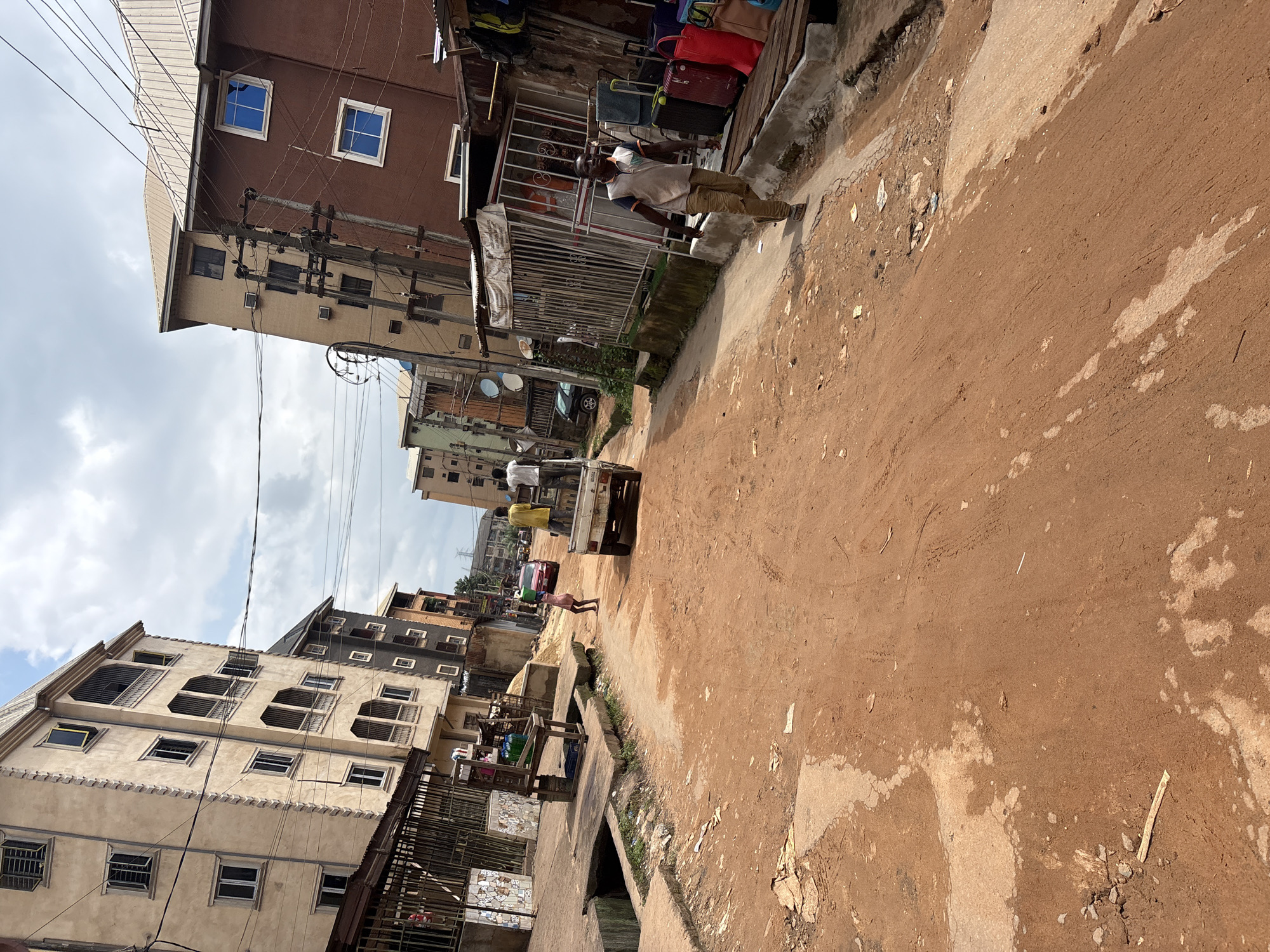 A street scene in Onitsha, Nigeria, features a reddish-brown dirt road with tire tracks and scattered debris. On the left, a tall, cream-colored apartment building with multiple floors and numerous windows, some with security bars, dominates the foreground. Adjacent to it, a lower structure with a metal fence encloses a space with various items, possibly for sale. Across the street, on the right, a brown apartment building with blue-framed windows stands. A utility pole with many wires is positioned between the buildings. Several people are present: a man in a light-colored shirt and tan pants walks on the right side of the road towards the viewer; two men stand in the back of a white pickup truck moving down the street; and a child in a pink dress walks ahead of the truck. Luggage and bags are stacked on the right side of the street near the brown building. The sky above is partly cloudy.