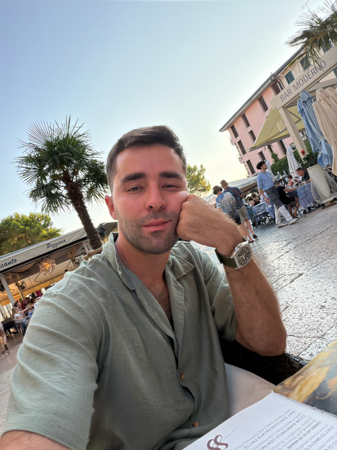 A man with dark hair and a beard takes a selfie outdoors, resting his chin on his hand. He wears a light green button-up shirt and a watch on his left wrist. In the background, a palm tree is visible, along with a pink building labeled "BAR MODERNO" and tables with umbrellas. The scene appears to be a cafe or restaurant located in Sirmione, Italy, on a sunny day.