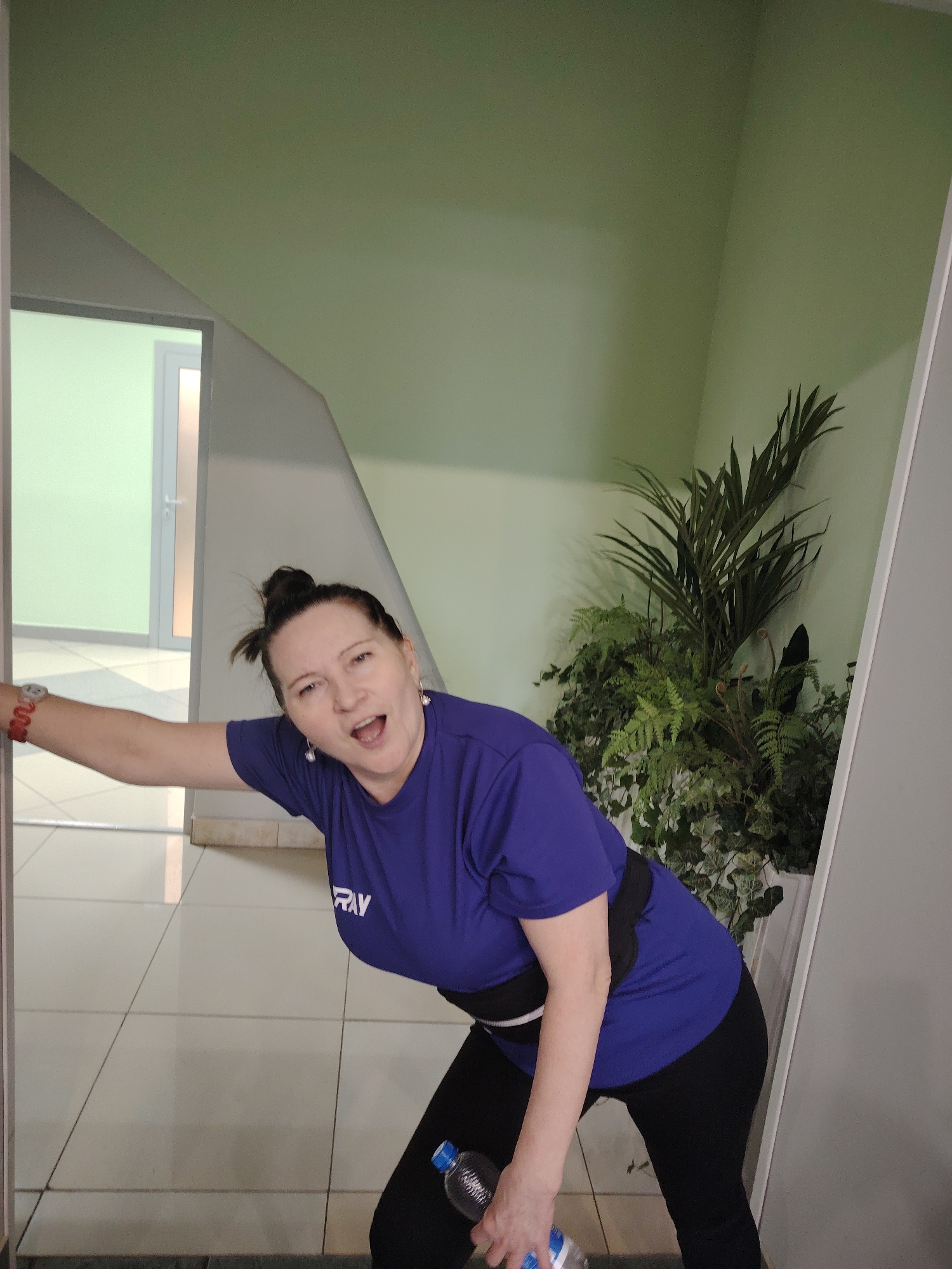 A middle-aged woman stands in a building in Sarapul, Russia, appearing in an entryway. She is wearing a dark blue shirt with white lettering and black leggings. A black and white belt encircles her waist. She holds a clear plastic water bottle in her right hand. Her left arm extends out towards a doorway. The woman's mouth is open in a possible expression of surprise or exertion. The floor is covered in square light-colored tiles. A potted fern and palm-like plant are visible to the right of the woman. The walls are a light green color. Another room is visible through the doorway to the left.
