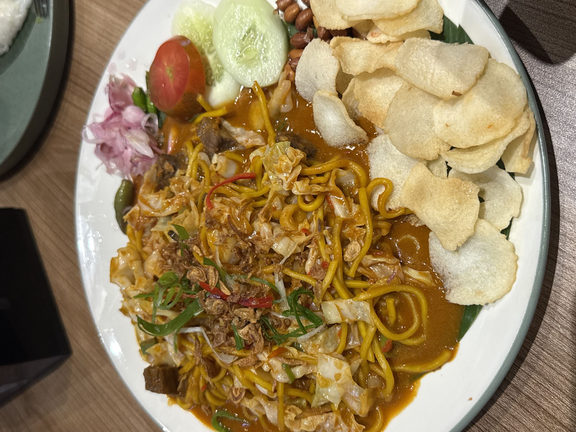 A close-up overhead shot shows a white plate of noodles with various accompaniments on a wooden table. The main dish is a mound of yellow noodles coated in a rich, orange sauce, mixed with shredded cabbage and topped with fried shallots and green onions. To the right of the noodles are slices of cucumber, a wedge of tomato, and a small pile of pickled red onions with a few green chilies. In the lower right corner, a scattering of peanuts and several light-colored crackers are visible. The plate has a subtle green rim. In the background, out of focus, a dark rectangular object is partially visible on the left, and a green plate with a fork is on the right. The lighting is dim, suggesting an indoor setting. There are no people or animals in the image. The scene is likely a restaurant in Gunung Putri, Indonesia, given the provided context.