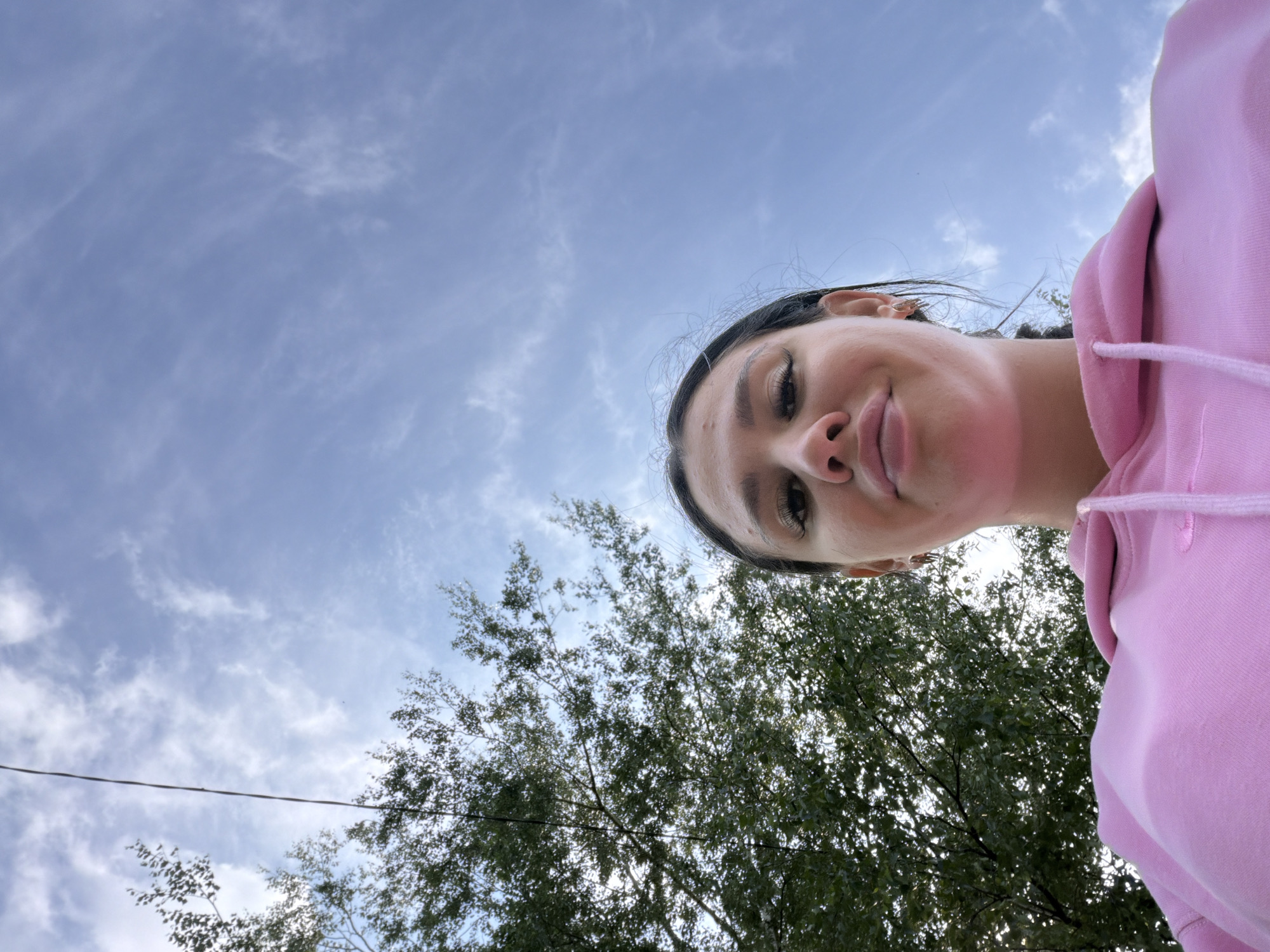 An outdoor, low-angle, selfie-style shot shows a young adult with dark hair and makeup, wearing a pink hoodie. The subject's face is partially visible from the chin up, with their head tilted back and looking towards the camera with a slight smile. Their eyes are looking up, and they have dramatic eye makeup, including false eyelashes. The background consists of a bright blue sky with scattered white clouds and the upper branches of a leafy green tree on the left side of the frame. A thin dark wire runs vertically through the upper left portion of the image. The location is described as Chumna, Russia.
