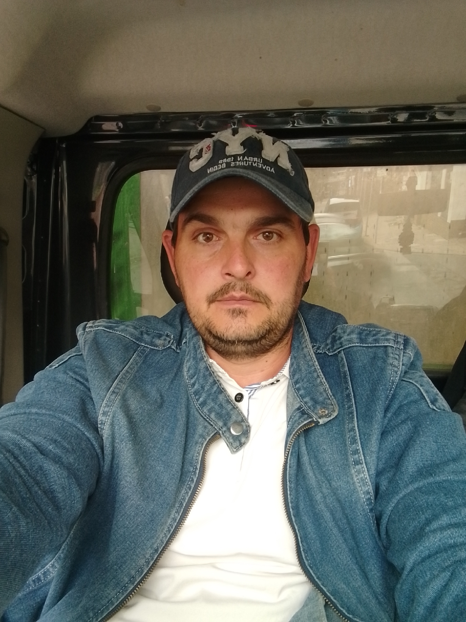 A man with a beard and brown eyes is sitting in a vehicle, looking directly at the camera. He is wearing a denim jacket over a white t-shirt and a baseball cap. The background is blurred, showing what appears to be an urban environment. There are no specific landmarks or features visible that would indicate the location as Pinneberg, Germany.