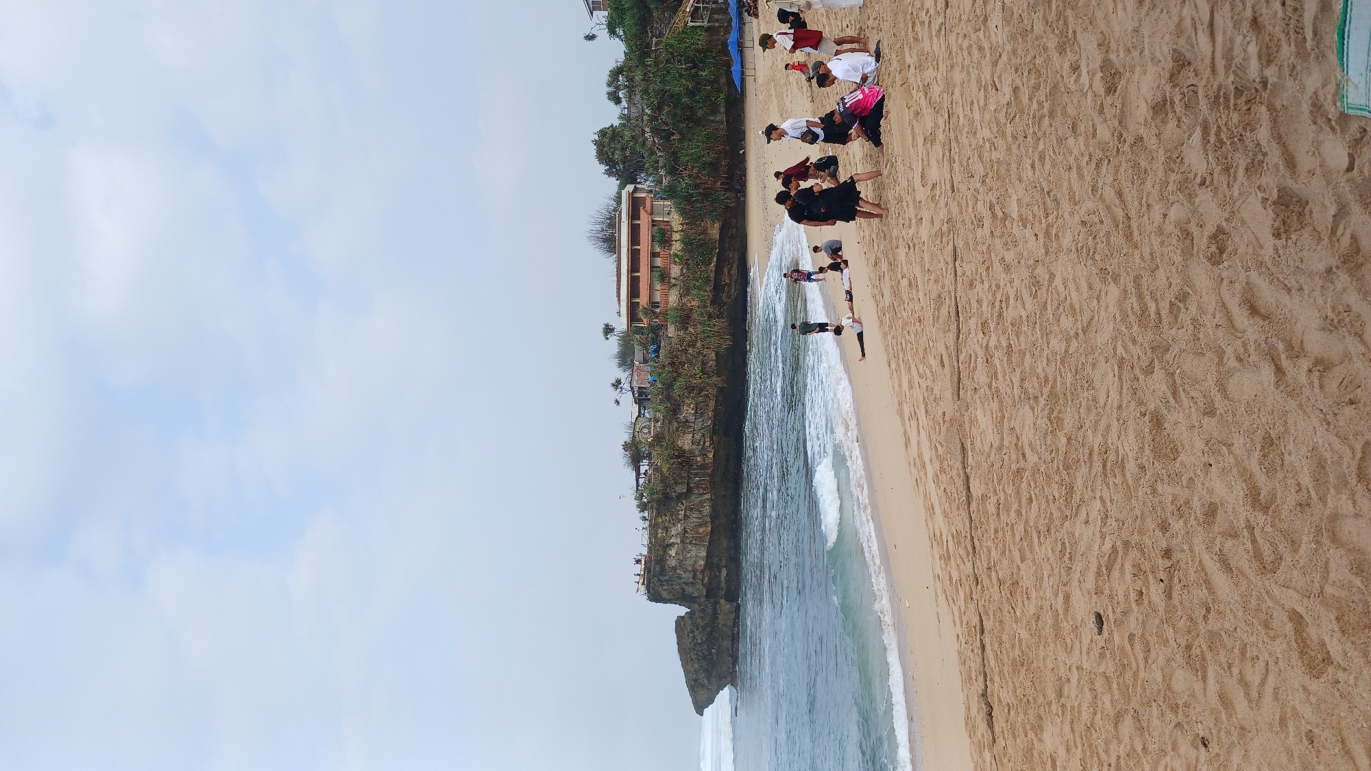 This is a vertical, eye-level, full shot of a crowded beach with a cliff in the background, taken in Tanjungsari, Indonesia. The sand is a light tan and has many footprints and some small debris. On the beach, there are at least 15 people, most of them young adults. Some are sitting in groups, some are standing, and one person is lying down. There is a body of water on the left, with waves breaking on the shore. In the background, there is a large, rocky cliff with greenery on top. There are also buildings constructed on top of the cliff, and some umbrellas are set up on the beach. The sky is cloudy and pale blue.