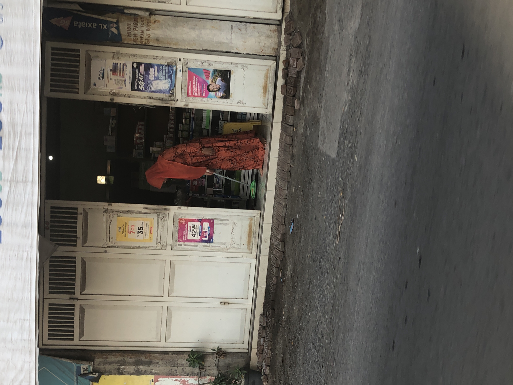 A person wearing an orange hijab and a patterned orange dress is sweeping the ground in front of a store entrance in Glagah Arum, Indonesia. The store has a white double door with vertical slats at the top. Advertisements are posted on the doors and the wall. The ground in front of the store is paved with asphalt and there are bricks lined up near the entrance.
