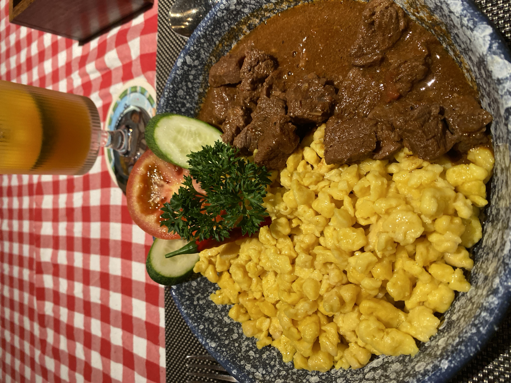 The image shows a plate of food, likely in a restaurant setting in Phu Thuy Ward, Vietnam. A glass of orange liquid stands on a red and white checkered tablecloth. The plate contains spaetzle, which are small, soft egg noodles, topped with a brown gravy and what appears to be pieces of beef. Garnishes include a slice of tomato, cucumber slices, and a sprig of parsley. The plate is set on a black woven placemat.
