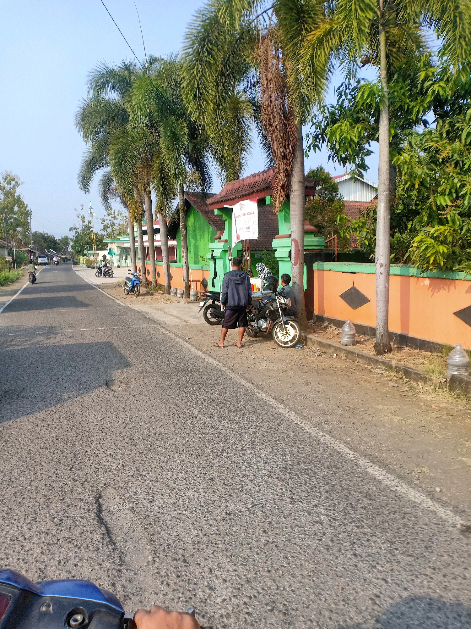 The image shows a road scene in Jatinom, Indonesia, with a focus on a building and surrounding elements. In the foreground, the textured asphalt road is visible, with a blue scooter part in the bottom left corner, suggesting the photo was taken from a moving vehicle. The road is lined with a dirt shoulder on the right.
Along the roadside, a vibrant orange wall with green trim borders a building. Several large palm trees stand tall in front of the building, casting shadows on the ground. Two men are present near a parked motorcycle. One man, wearing a dark hooded jacket and shorts, stands facing away from the camera, near a motorcycle with a black seat. Another person is seated on the motorcycle. A second motorcycle, blue and white, is parked further down the road on the shoulder.
Further down the road, other vehicles and buildings can be seen. In the distance, there are more palm trees, foliage, and what appears to be a water tower or tall structure with antennas. The sky is clear blue, indicating a sunny day. The overall scene depicts a typical street in a residential or semi-commercial area in Jatinom, Indonesia.