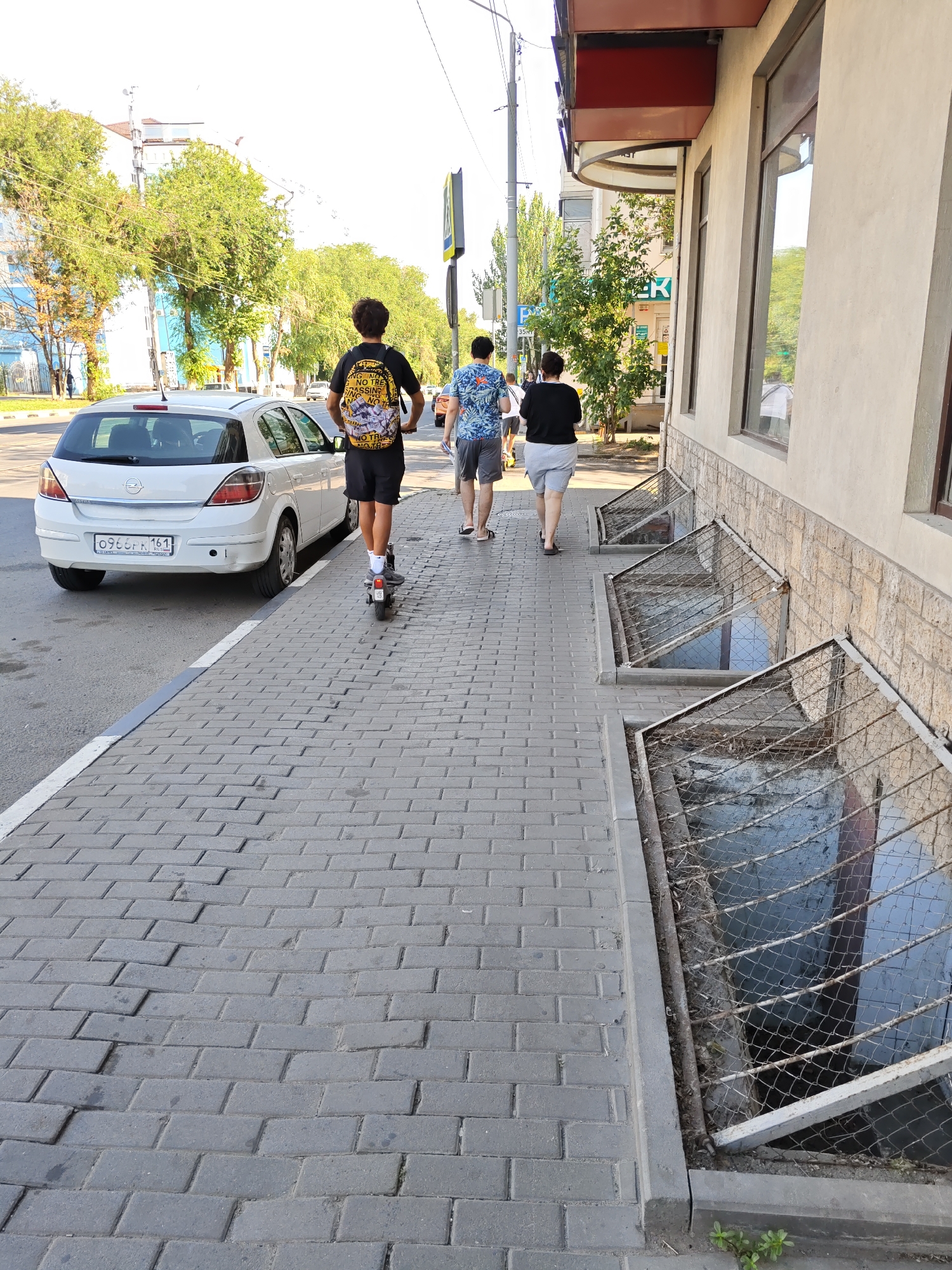 A young man on an electric scooter, wearing a black t-shirt with a yellow graphic and black shorts, travels along a gray brick sidewalk. He is followed by two young men, one in a blue floral shirt and black shorts, the other in a black t-shirt and gray shorts. They are walking away from the viewer. To their left is a white hatchback car parked on the street. The sidewalk is adjacent to the street and a building with a light-colored facade and windows. The building has several window wells covered with metal grates. The sky is bright, indicating daylight. The scene appears to be in Rostov-on-Don, Russia, based on the license plate of the car, which reads "0966PK 161". The overall environment is urban, with trees visible on the left side of the street and buildings in the background.